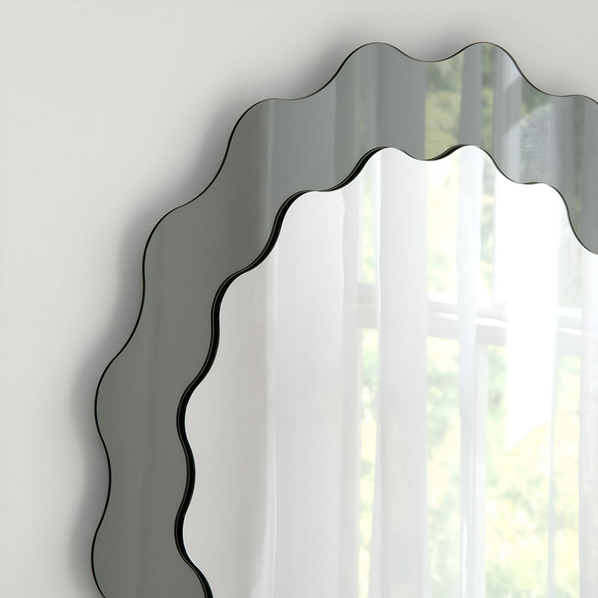 The Solstice Circle Wall Mirror - Grey, with its sculptural artistry and scalloped edge, reflects sheer white curtains and soft greenery while mounted on a light wall, adding a serene, minimalist touch to the space.