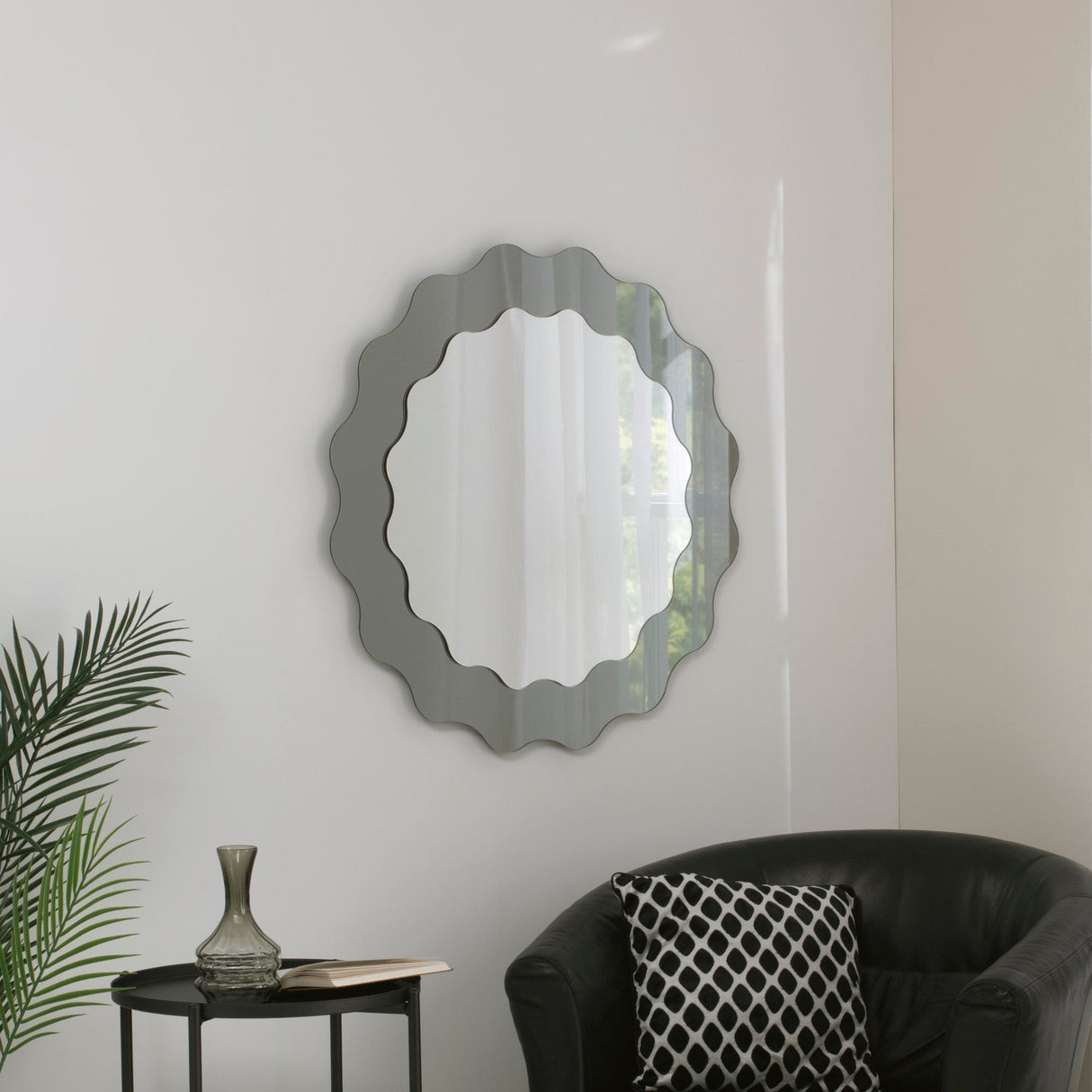 The Solstice Circle Wall Mirror - Grey with wavy edges hangs on a white wall above a black table with a glass vase, next to a black armchair featuring a geometric pillow and a nearby potted plant.