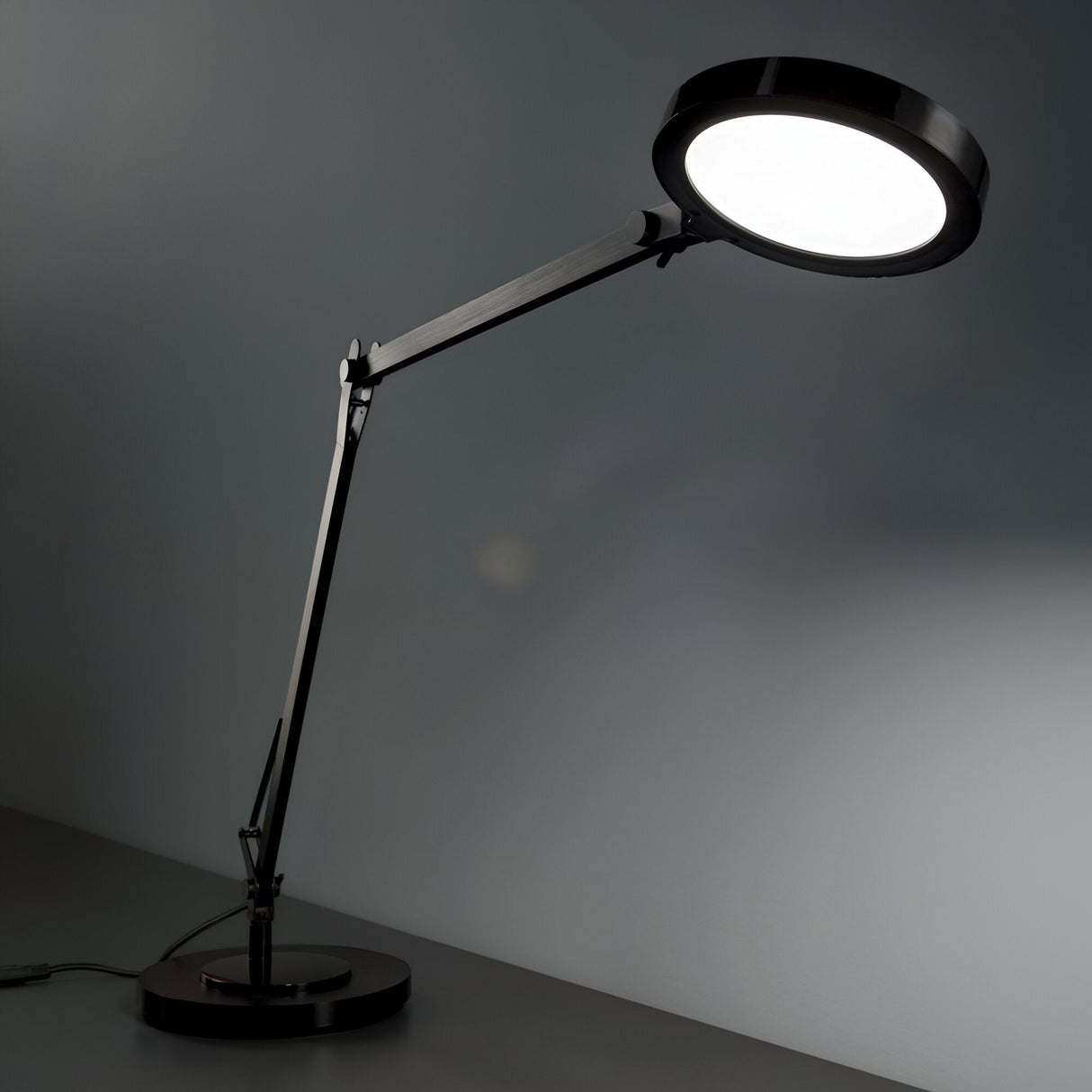 The Prismora LED Table Lamp 12.5W 4000K in black presents an industrial-inspired glow with its sleek round light head over the surface. Its articulated arm and adjustable LED diffuser provide flexible positioning against a plain, gray background.