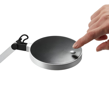A hand touches a sleek, silver Prismora LED Table Lamp, characterized by its modern design and round black base, with an adjustable arm. The background is white.