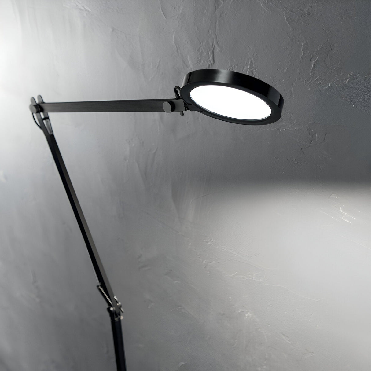 The Prismora LED Floor Lamp 12.5W 4000K in black, featuring a circular light head and minimalist design, illuminates a textured gray wall. With its flexible positioning, the lamp extends diagonally from the left side of the image, casting a soft glow on the surface.