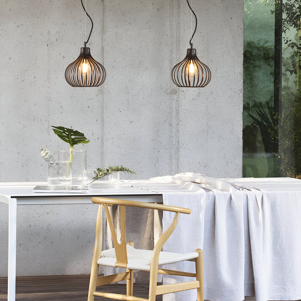 A modern interior boasts the stylish Solyra 1 Light Pendant in brown (23cm) elegantly hanging above a white table. A wooden chair is positioned next to it, decorated with glass vases and leaves. Through a large window, greenery outside enhances the space's contemporary elegance.