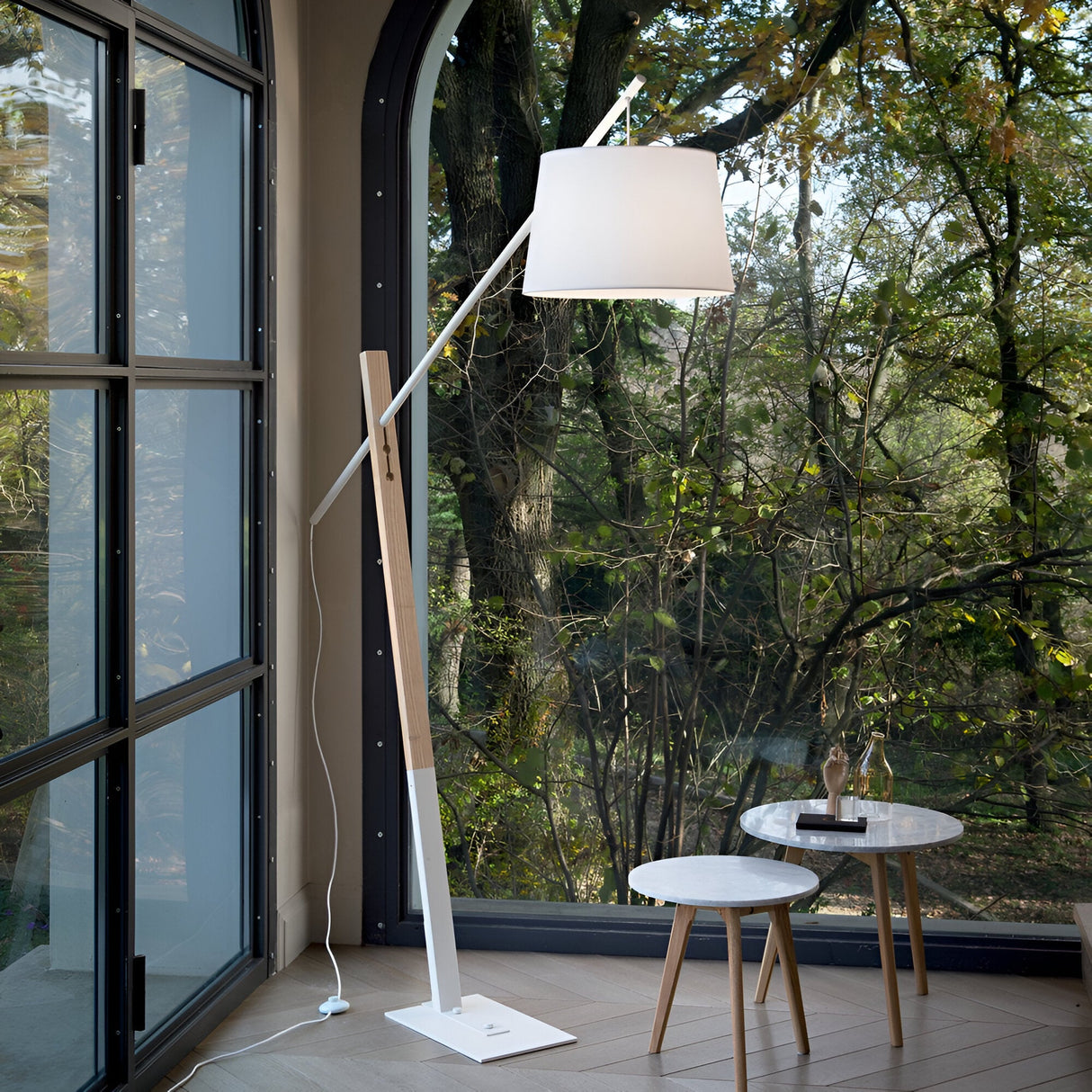 A modern living room exudes natural elegance with the Solixia Wood Floor Lamp, featuring a white shade, positioned next to large arched windows. The view outside reveals a dense forest. Nearby, a small round table with two stools holds a decorative item and a book, embodying modern simplicity.