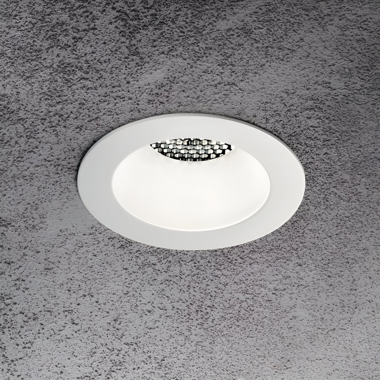 A circular Lumos Downlight Round Frame - White, with a modern design, is installed on a textured grey ceiling. This recessed ceiling light offers ambient lighting and includes decorative elements inside its white rim.