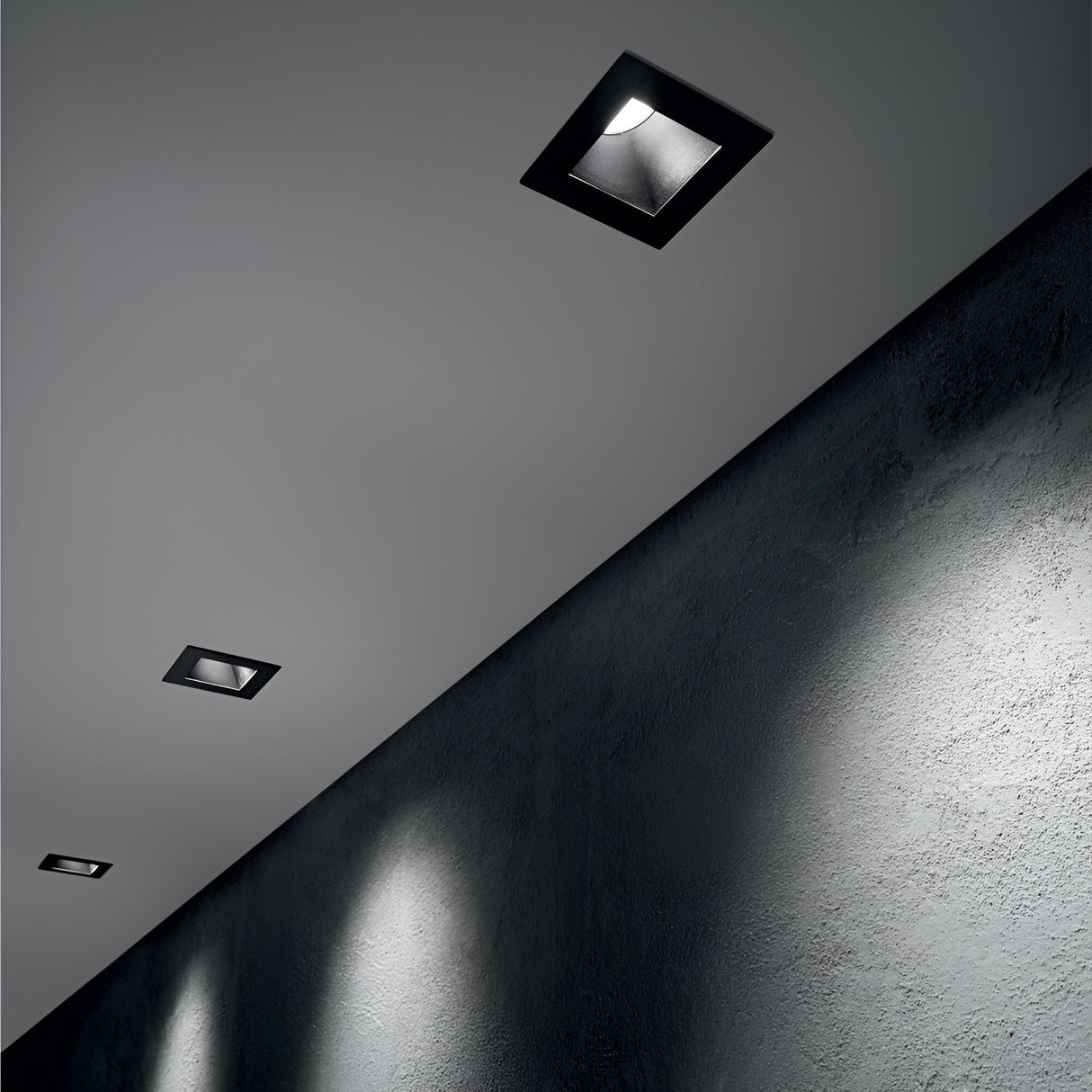A sleek, modern interior ceiling showcases three Lumos Downlight Square Frame fixtures in black elegantly recessed, casting soft glows on a textured gray wall below. The minimalist design features modular light sources that create a calming ambiance.