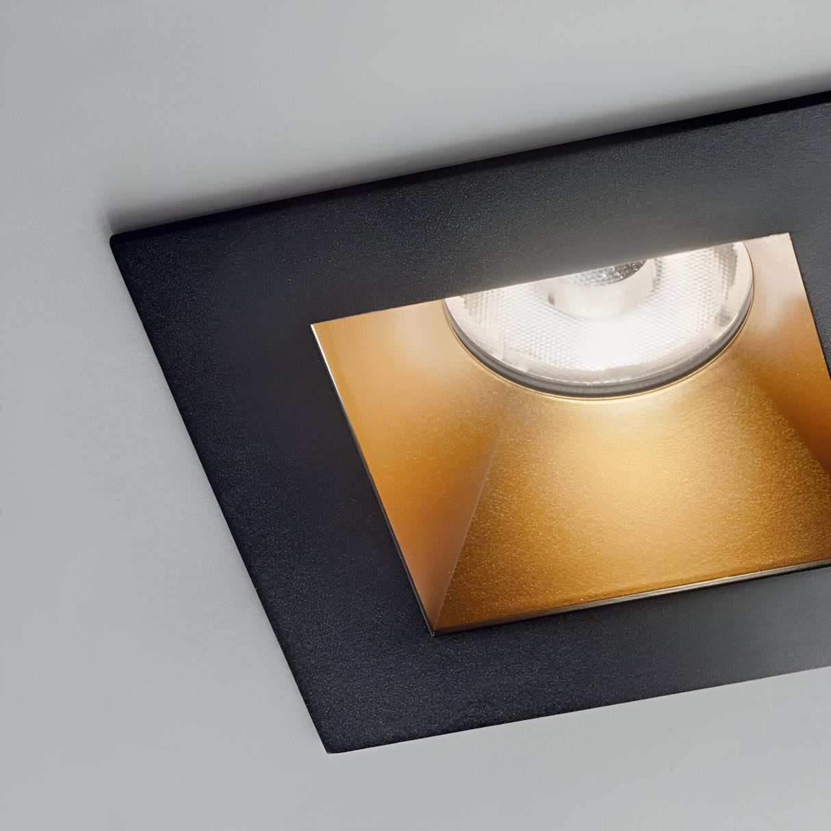 Close-up of the Lumos Downlight Square Frame - Black, highlighting its black square frame paired with a gold reflector. The recessed light casts a bright glow, emphasizing the modern and sleek design of this modular light source against a white ceiling.
