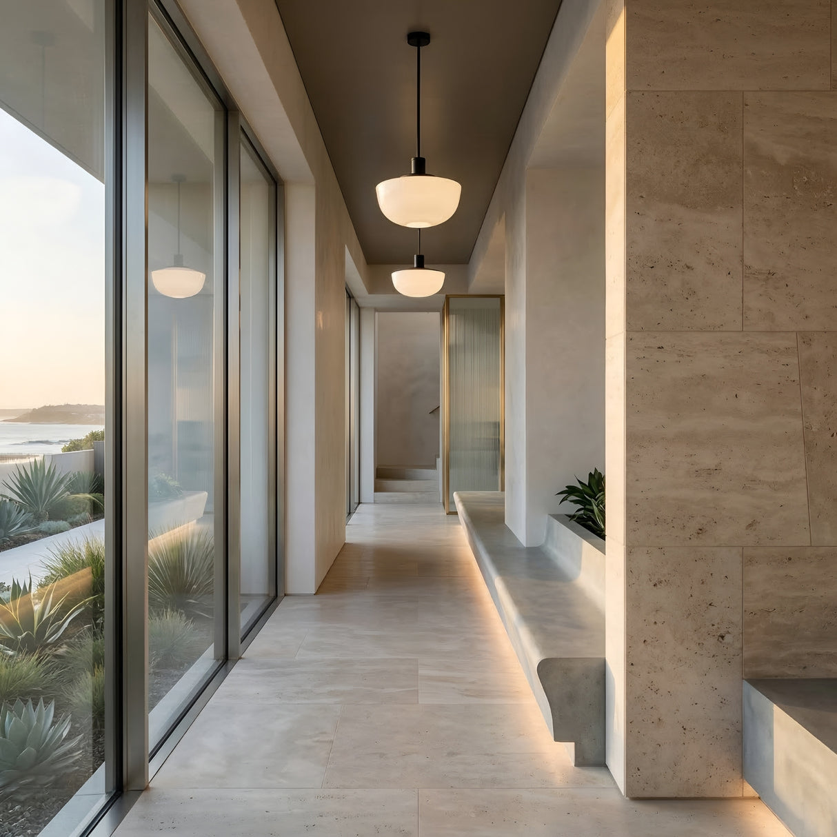 A modern hallway with stone tile, built-in bench seating, and large floor-to-ceiling windows showcases Verge 1 Light Pendant - Opal Shade fixtures, offering views of a sunlit desert landscape with plants.