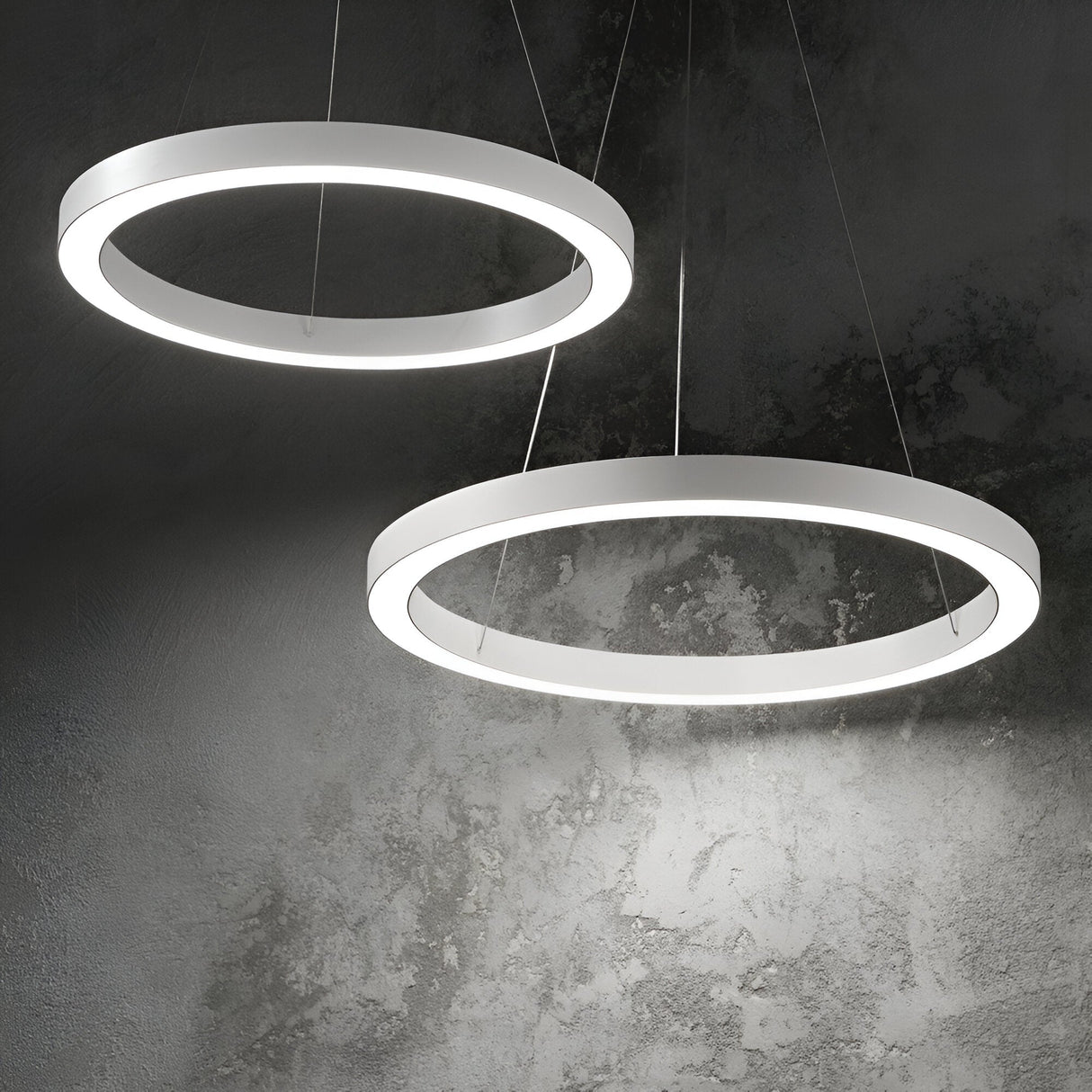 Two Arcena Round LED Suspension Lights, each featuring a glowing ring, are mounted against a textured gray wall, casting soft light and subtle shadows. The sleek and minimalist design of this dimmable, energy-efficient fixture creates a contemporary ambiance.