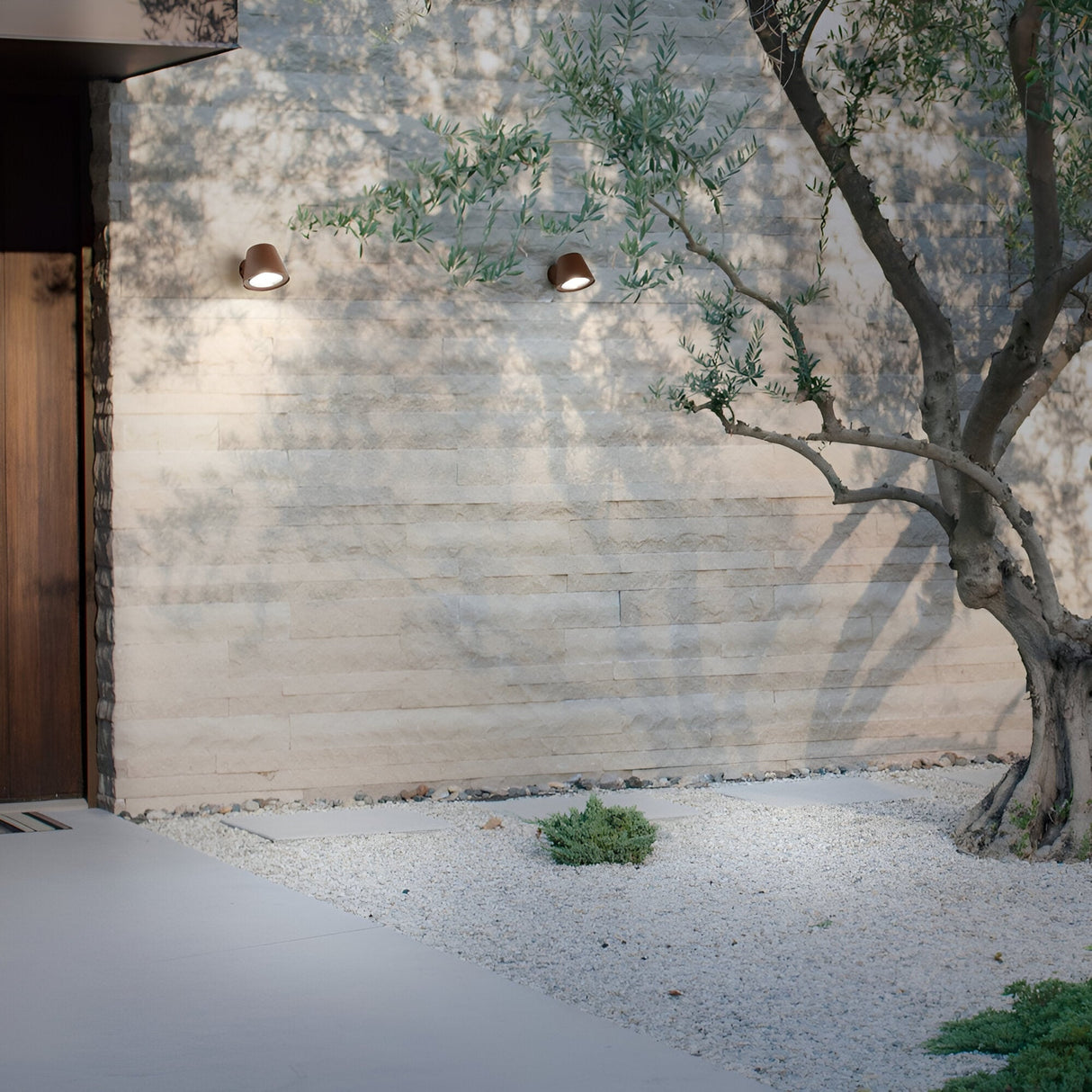 A minimalist outdoor space showcases a sand-colored stone wall adorned with two Quasar Outdoor Wall Lights in brown. An olive tree casts shadows on the gravel ground, which is dotted with sparse green plants that add a rustic touch. A wooden door is partially visible on the left.