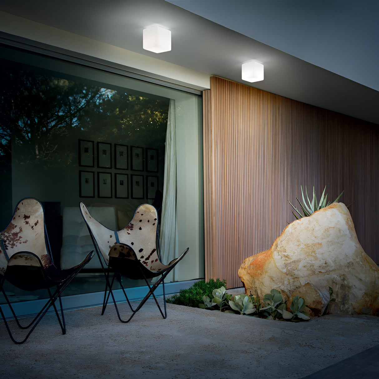 In a contemporary patio setting, two stylish chairs are positioned to face a large glass window, with vertical wooden slats adorning the wall and a sizable rock accompanied by plants on the right. The chic space is illuminated by two Spectra Outdoor Ceiling Lights in white, each with an opal diffuser, measuring 20cm in diameter.