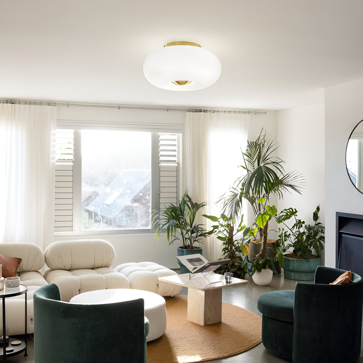 A modern living room with a Solitha 5 Light Flush - White ceiling fixture. The space features a white sectional sofa, green velvet chairs, and a marble coffee table. Large windows with sheer curtains welcome natural light, while potted plants add greenery.