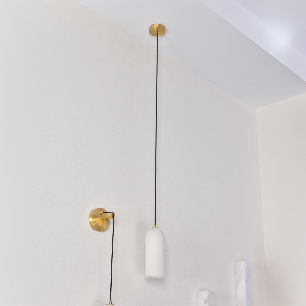 Velmora slim cylindrical alabaster pendant light in soft white, with black flex cable and brass ceiling plate, shown alongside a matching brass wall light in a bright interior.