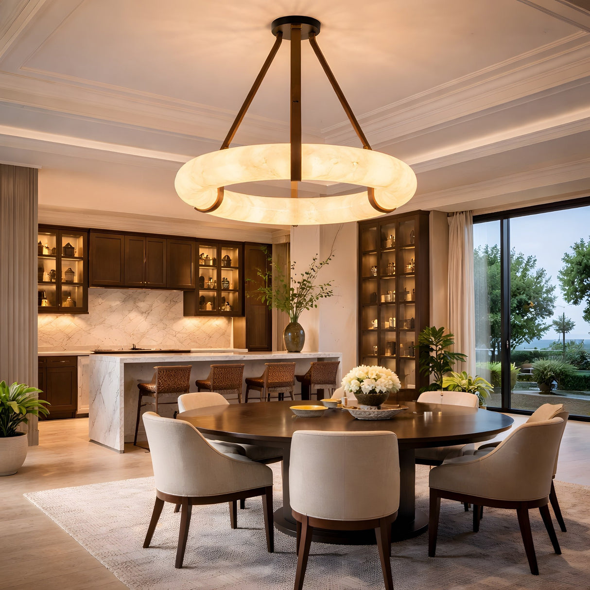 A modern dining room features a round table, six upholstered chairs, and an open kitchen with dark cabinets and marble surfaces. The Cintura LED Medium Alabaster Suspension Light in soft white & brown hangs under floor-to-ceiling windows overlooking greenery.