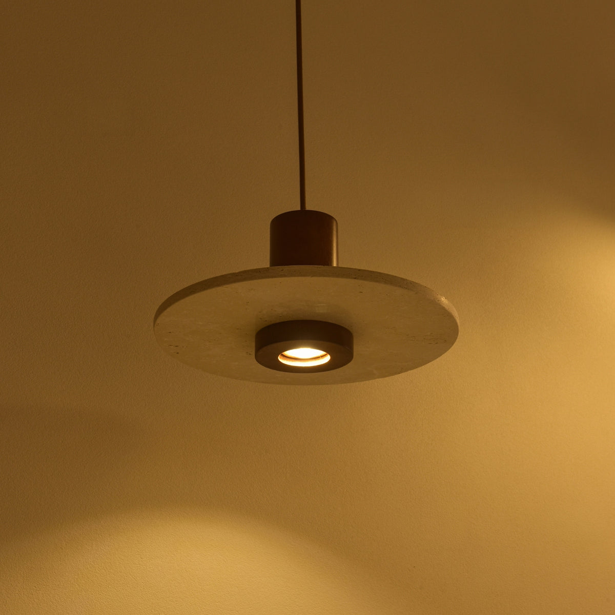 Torvane 1 Light Travertine Disc Pendant Light with light beige stone disc, dark walnut brown wooden column, and recessed downward-facing light, shown illuminated against a warm wall.
