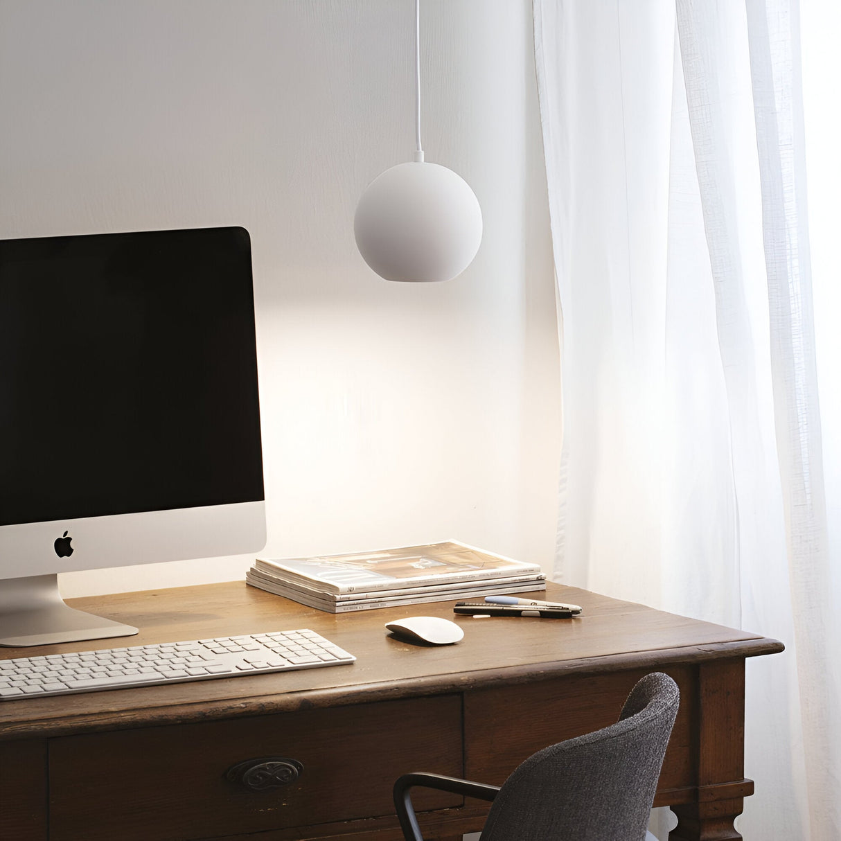A minimalist workspace featuring a wooden desk with an iMac computer, wireless keyboard, and mouse. Stacked magazines and pens accompany the Emberleaf Large 1 Light Pendant - White, whose contemporary lighting complements the setup. Sheer white curtains allow soft natural light into the room.