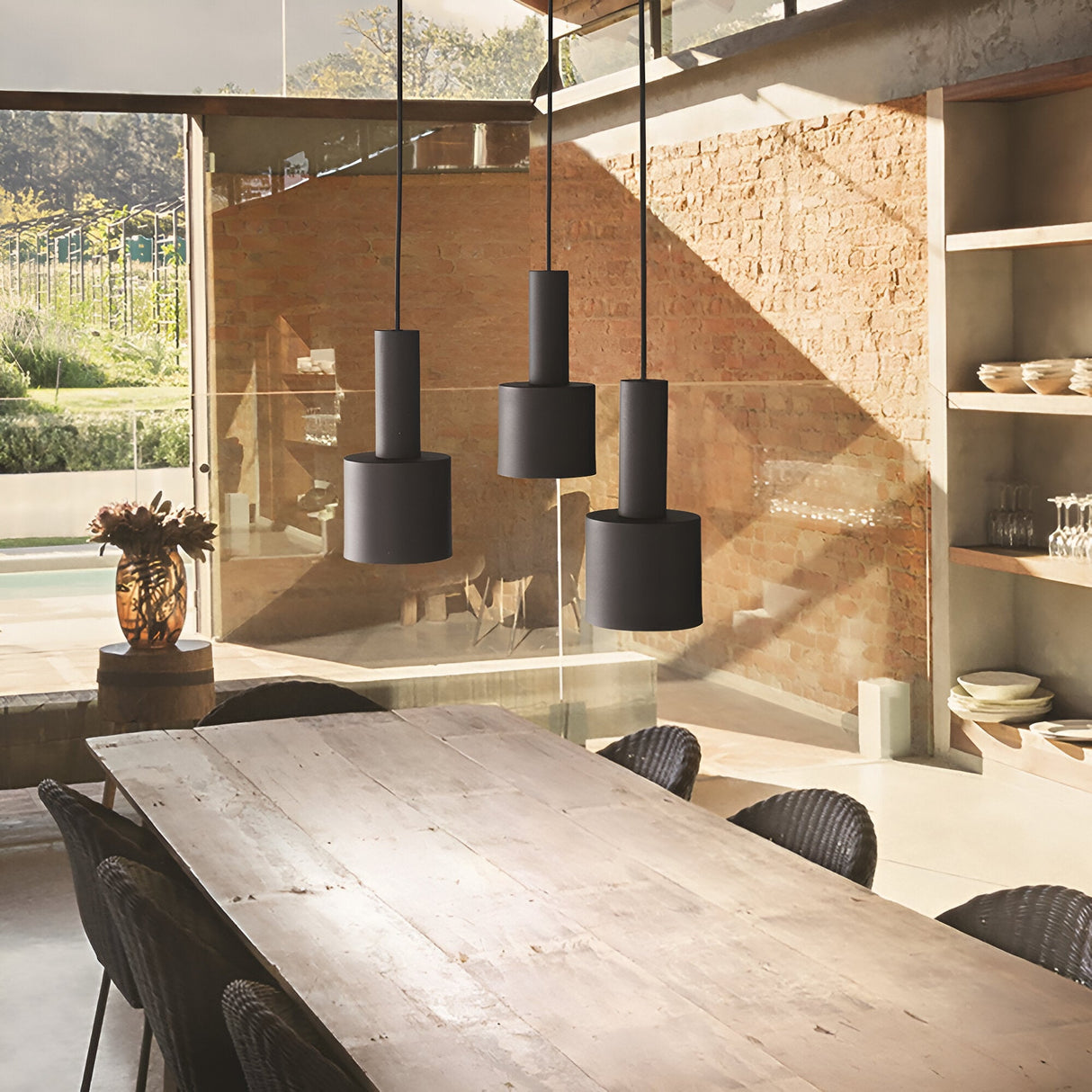 A modern dining area showcases a wooden table and wicker chairs beneath the Astro 1 Light Pendant - Black, creating a sleek lighting effect. The glass wall offers views of the garden and pool, while exposed brick and open shelves with plates enhance the minimalist charm.