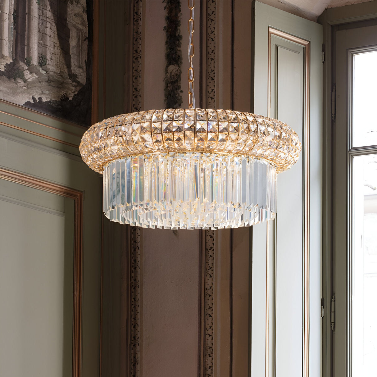 The Enluma Crystal 12 Light Chandelier in gold boasts a luxurious design with two tiers of pendants, elegantly suspended from a chain. Its warm glow beautifully enhances the intricate details of the ornate walls and large windows in the sophisticated room.