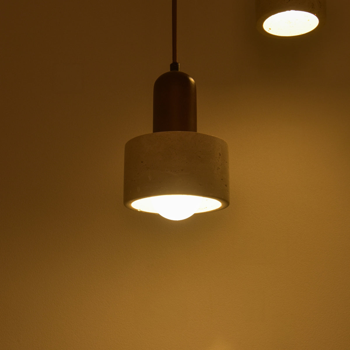 Lunaro 1 Light Travertine Pendant Light with cylindrical light beige stone shade and walnut wood upper section, lit and hanging on a brown cable in a warm-toned room.