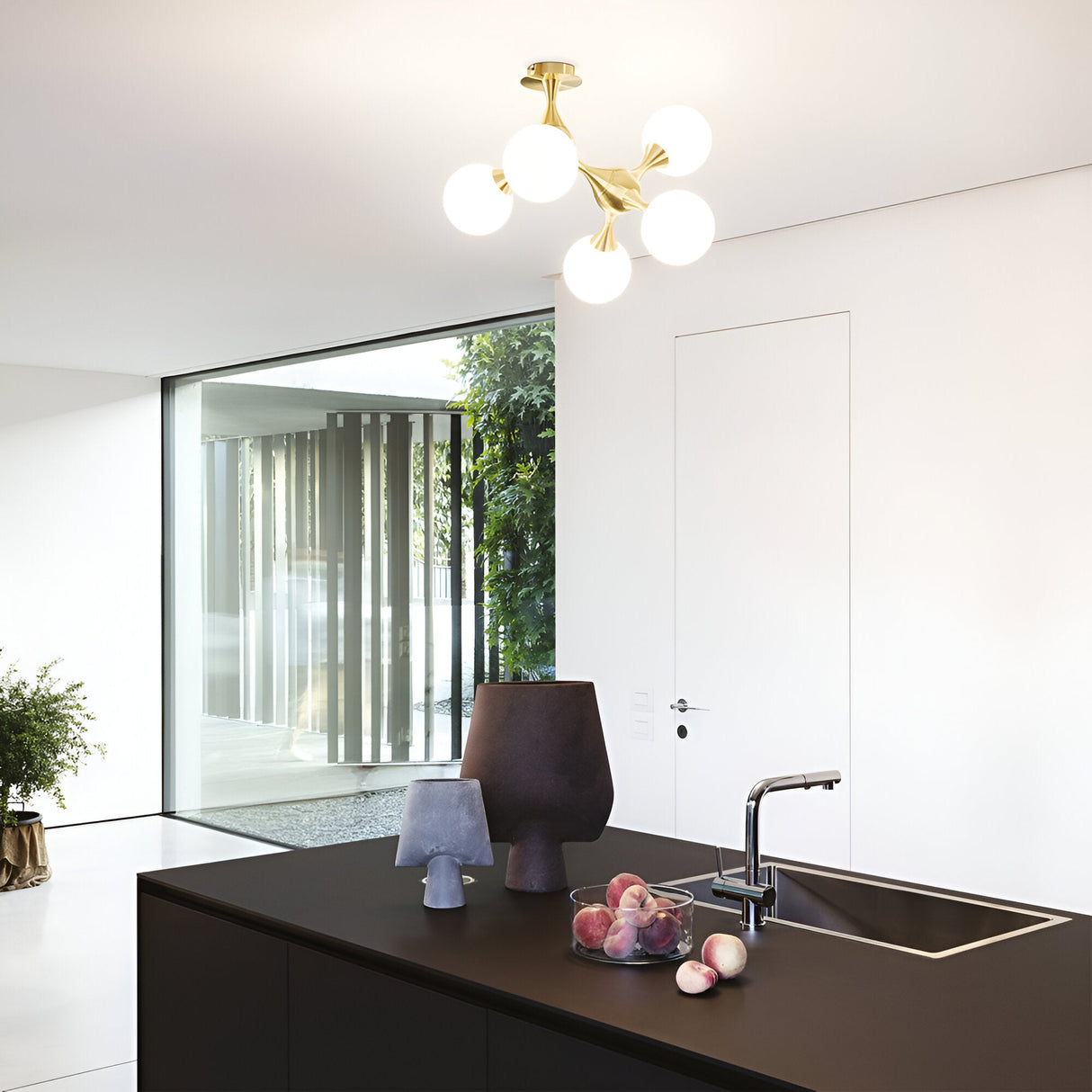 A minimalist kitchen showcases a sleek black island with a modern faucet, complemented by a trio of decorative vases and a bowl of peaches. Overhead, the Emblaze 5 Light Semi Flush in brass adds style with its frosted glass shades. Large windows on the left flood the space with natural light, offering views of greenery outside.