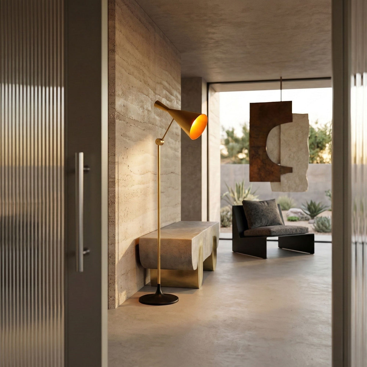 A modern, minimalist living room with concrete walls and floors, a Funk Floor Lamp - Brass, abstract wall art, and a black sofa by large windows overlooking a desert landscape.