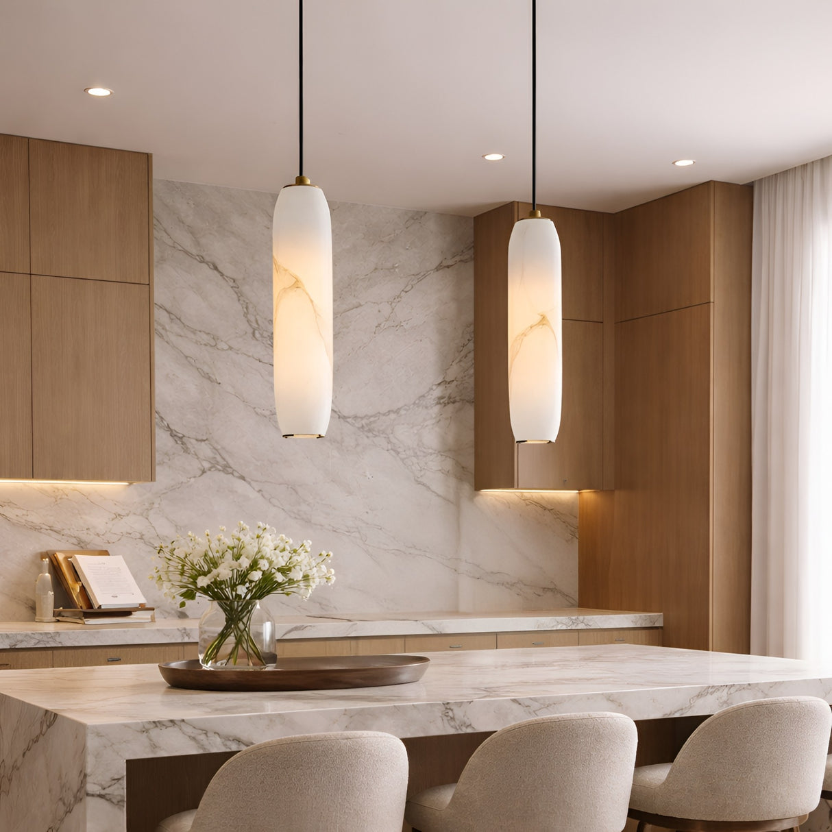 Modern kitchen with light wood cabinets, marble backsplash and countertops, Vireth 1 Light Small Cylinder Alabaster Single Pendant Lights in soft white, beige upholstered chairs, and a vase of white flowers on the island. Natural light comes through sheer curtains.