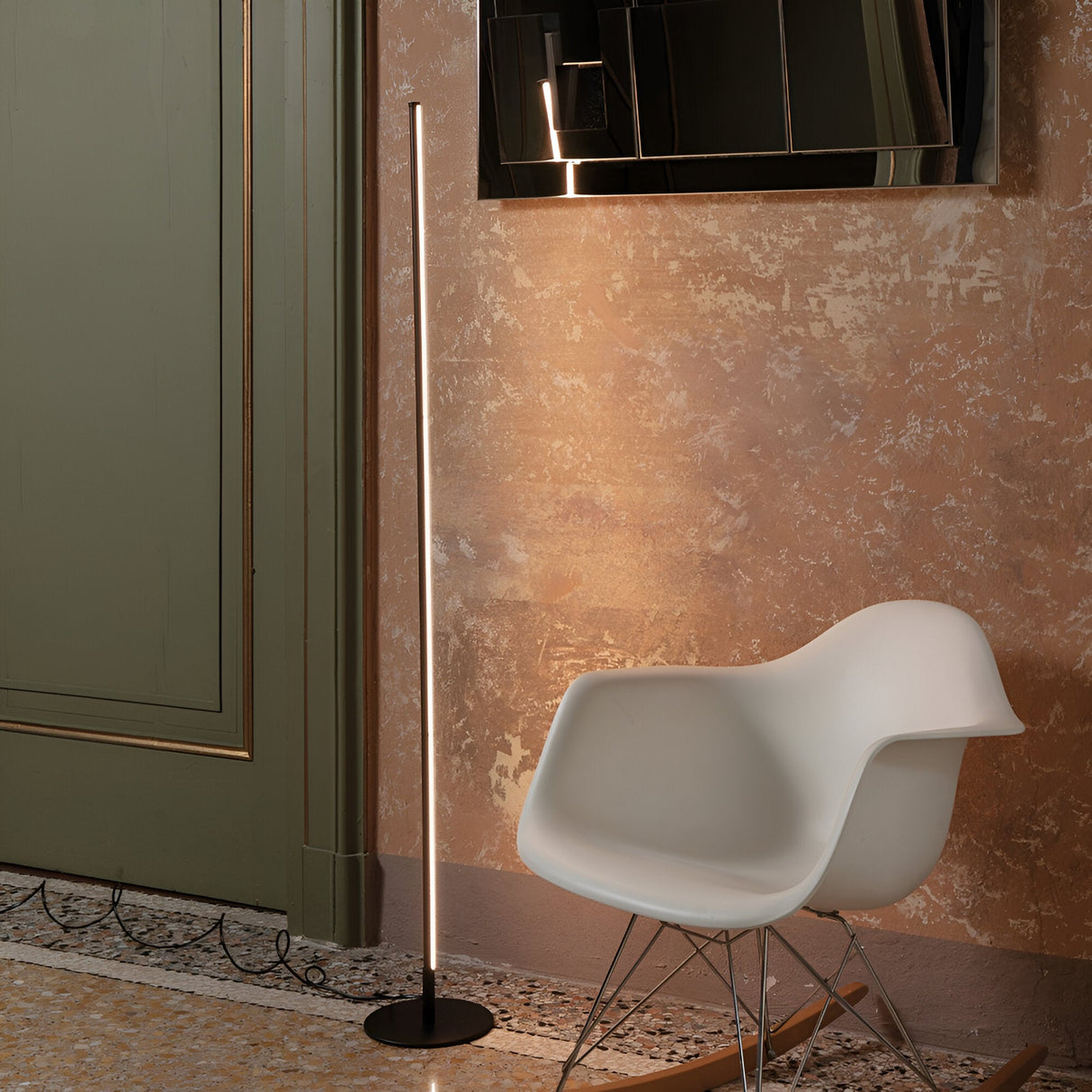 In a contemporary interior, the Wave LED Floor Lamp 16W 3000K in black casts a warm glow on a textured wall. Nearby, a white molded chair rests on the tiled floor. Above it all, a black-framed mirror reflects the room's modern lighting, adding depth to the decor.