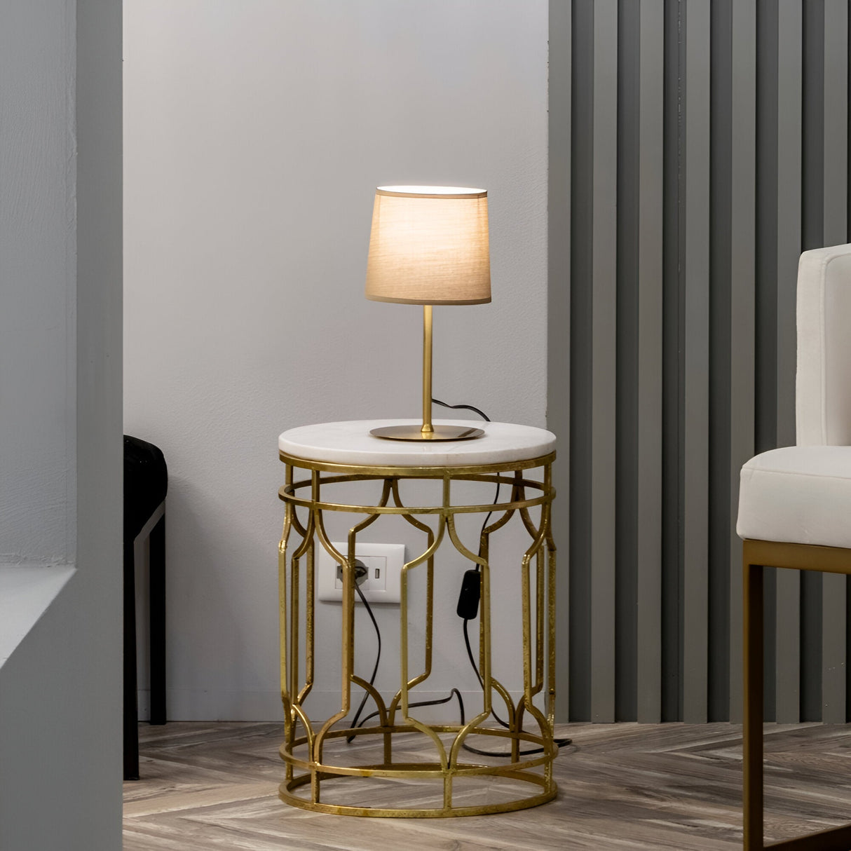The Aethera Small Table Lamp - Brass, complete with a beige shade, is elegantly positioned on a round white marble table adorned with a brass finish. This sophisticated arrangement enhances the modern room's aesthetic, harmonizing with the gray walls, herringbone-patterned wood flooring, and tall chair in proximity, creating an ideal home decor setup.