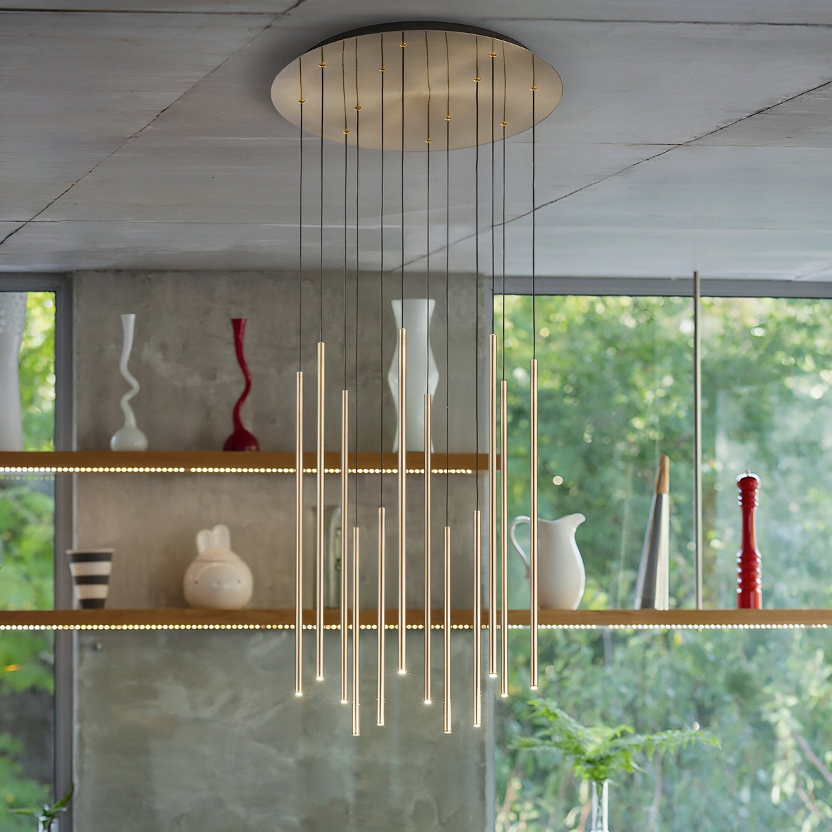 The Celestia 12 Light LED Cluster Pendant 25W 3000K - Brass illuminates the space with its minimalist design, showcasing vertical tube lights suspended from a round ceiling fixture. In the background, shelves adorned with assorted vases and sculptures are set against large windows that overlook lush greenery.