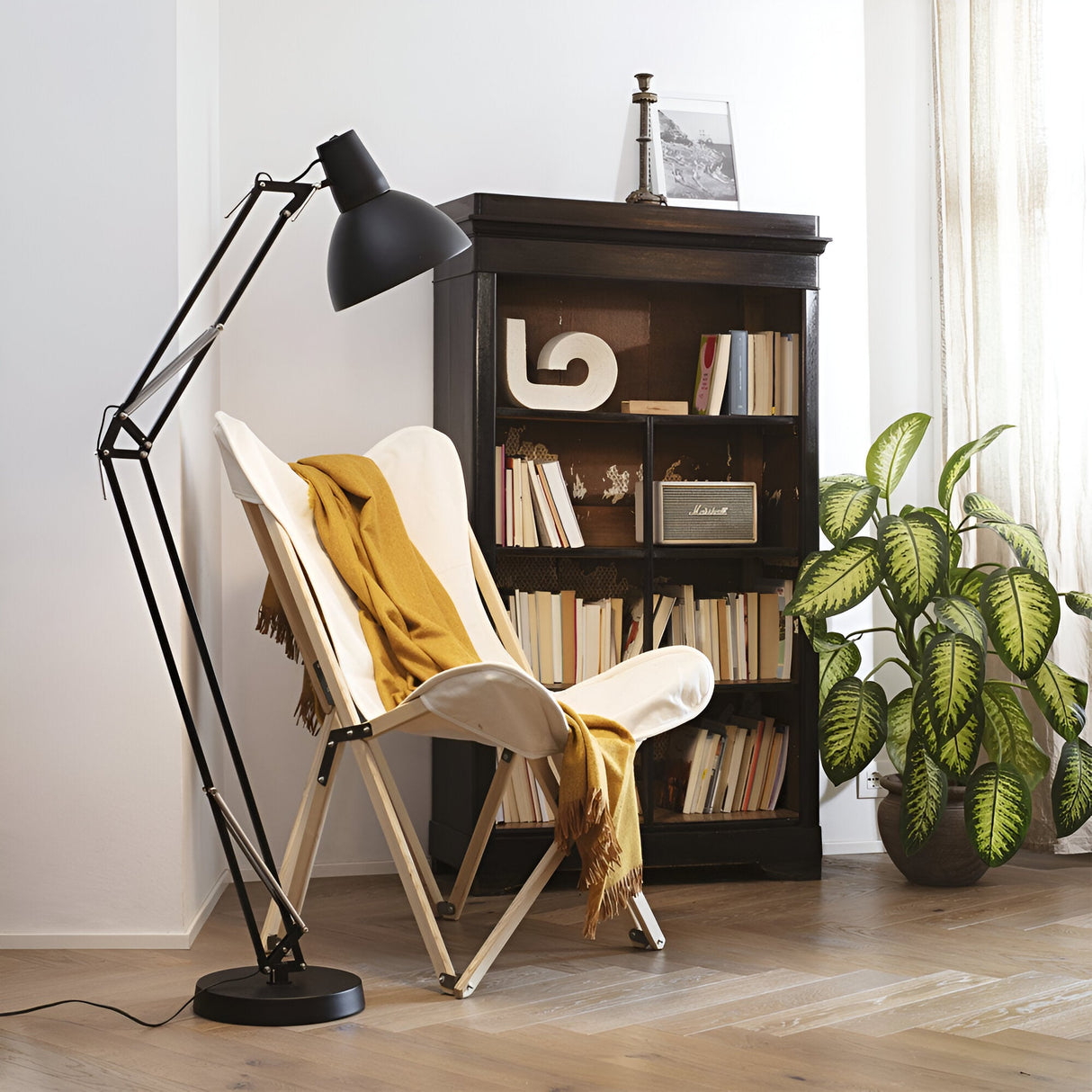 A cozy reading nook features a white butterfly chair draped with a mustard-yellow throw, illuminated by the sleek Maive Floor Lamp in black, featuring an adjustable arm. An industrial-style, dark wooden bookshelf filled with books stands nearby, complemented by a green potted plant enhancing the serene atmosphere.