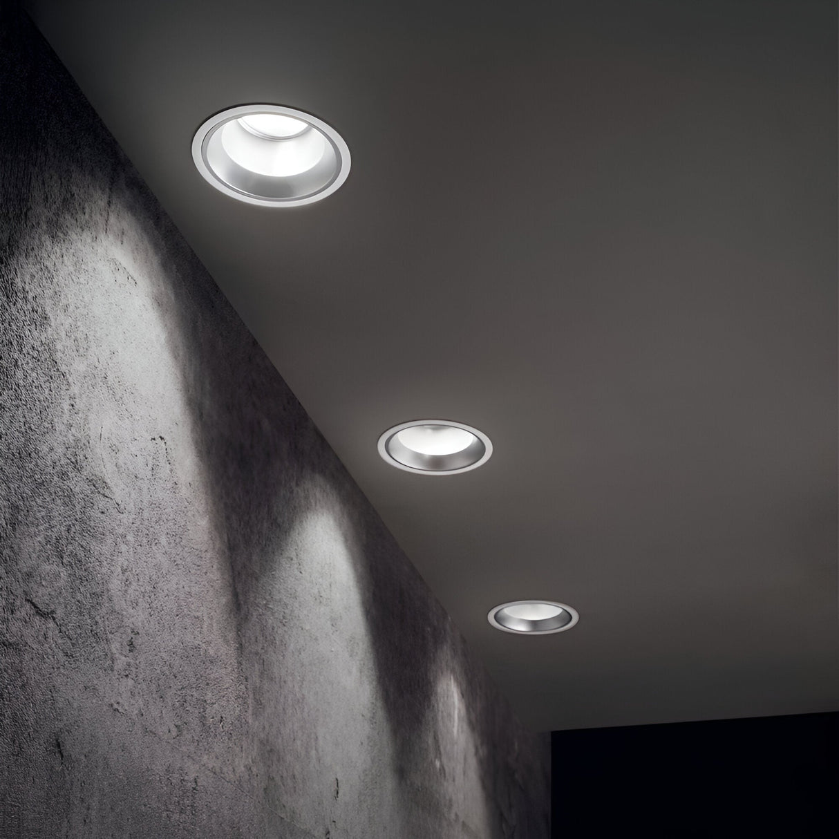 A ceiling featuring three Zeal Outdoor LED Recessed Downlights, each with a 28W luminaire and an IP54 rating, illuminates the textured dark gray wall with a subtle, focused glow. The evenly spaced arrangement of these lights establishes a modern and minimalist ambiance suitable for both indoor and outdoor environments.