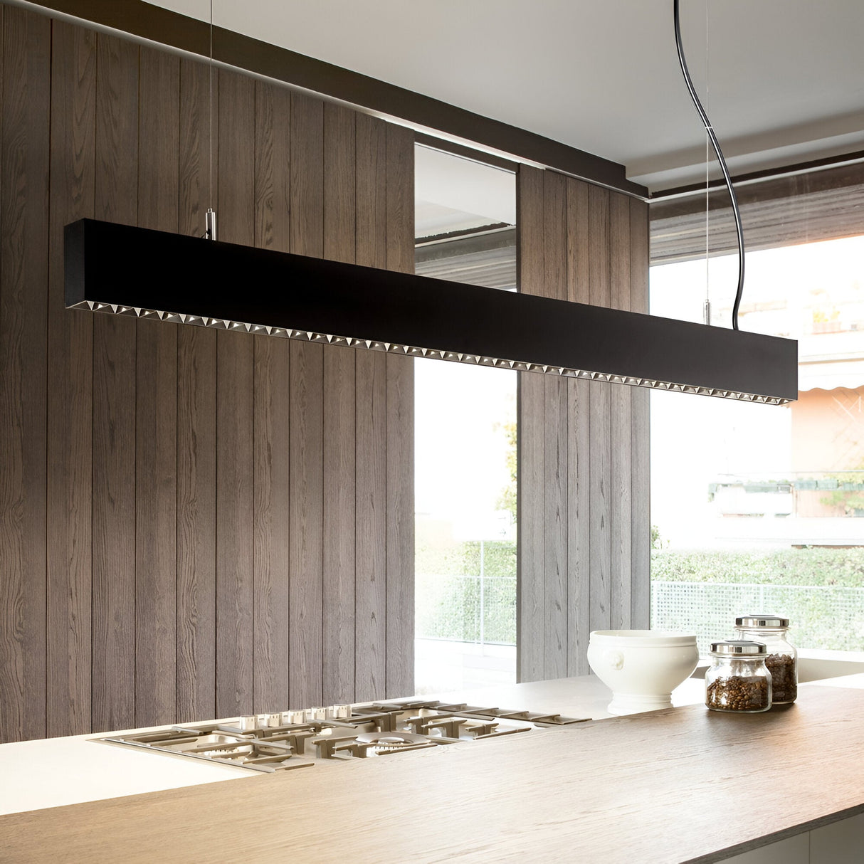In a modern kitchen, the Ignitia-2 LED Suspension Light 36W 3000K in black hangs above the light wood island, showcasing its sleek rectangular design with a matte black finish. This contemporary-style lighting elegantly illuminates the built-in stovetop, while white bowls and jars enhance the wooden paneling and large window in the background.