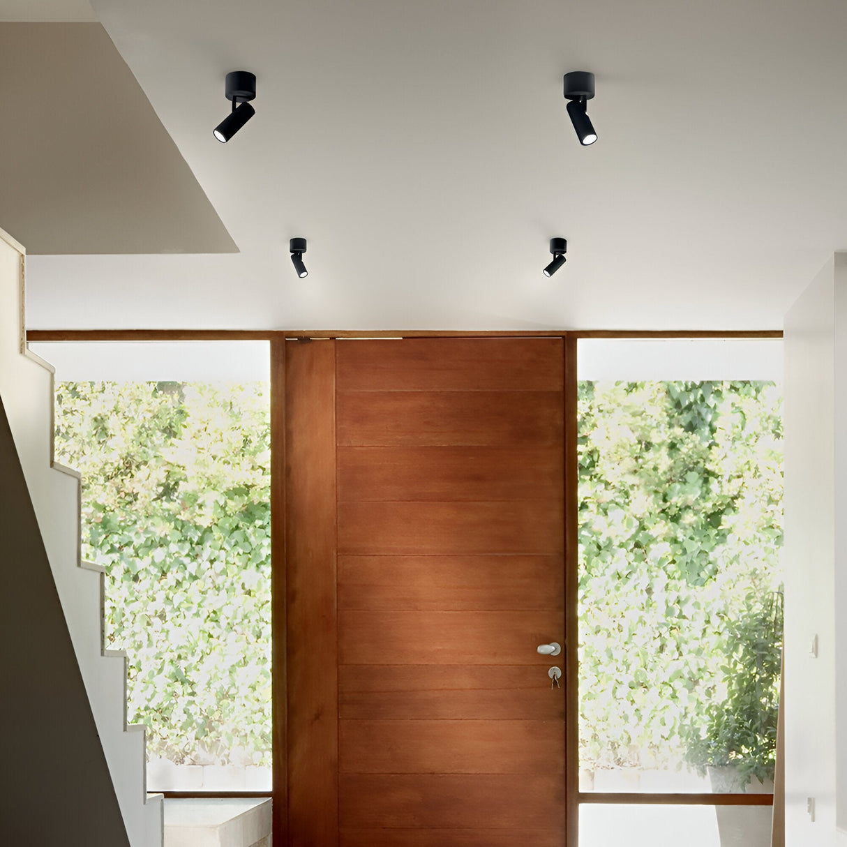 The modern interior includes a wooden door, white walls, and a staircase with white steps. The ceiling is equipped with three Opal LED Single Spotlights 7W 3000K in black, providing precise lighting. A large window allows views of the greenery outside, resulting in a minimalist and well-lit atmosphere.