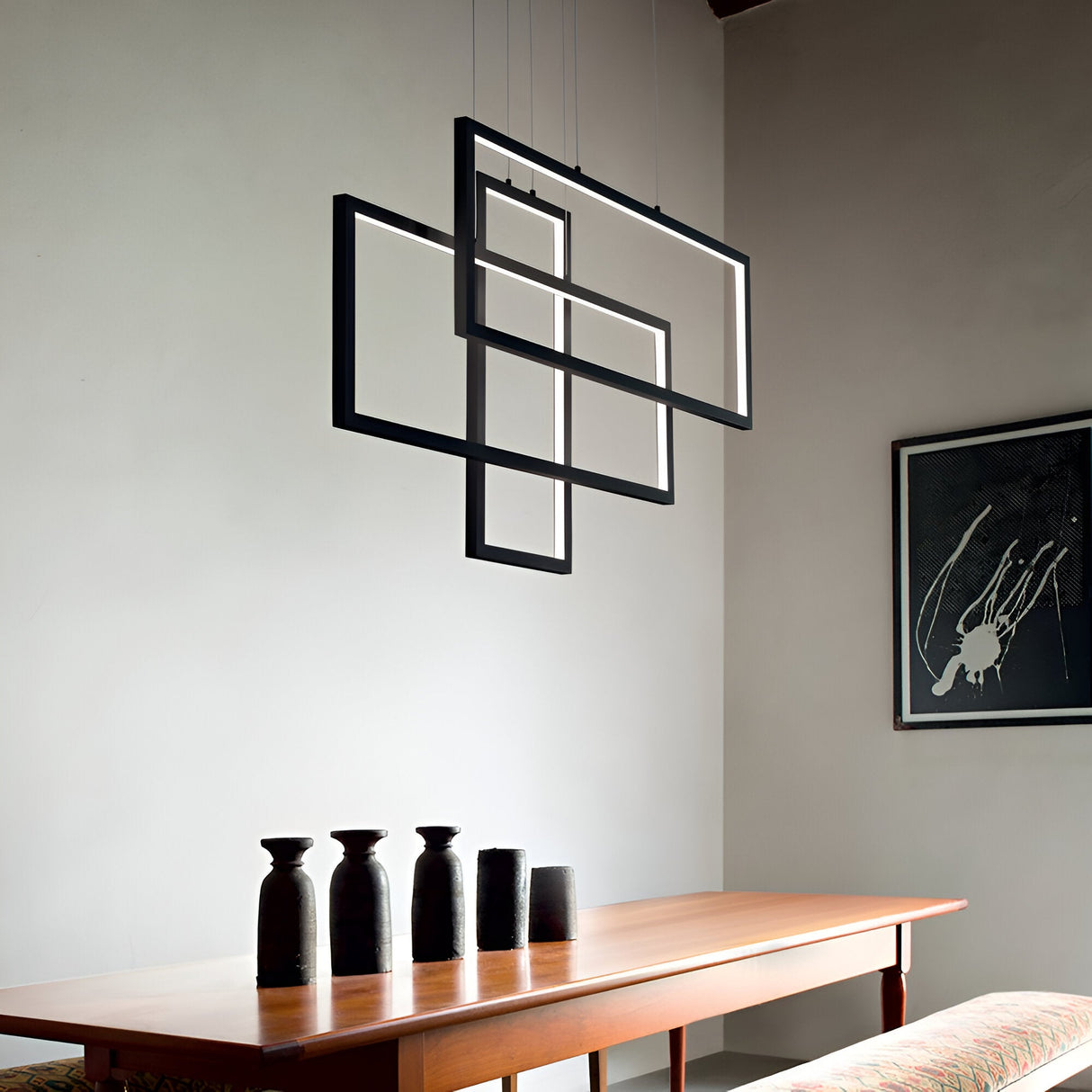A minimalist dining room is centered around a wooden table adorned with four black vases, while the Prios Rectangle LED Light Suspension 93W 3000K in Black, featuring an artistic geometric design, hangs elegantly above. A modern abstract black and white painting graces the wall, enhancing the sleek and contemporary atmosphere.