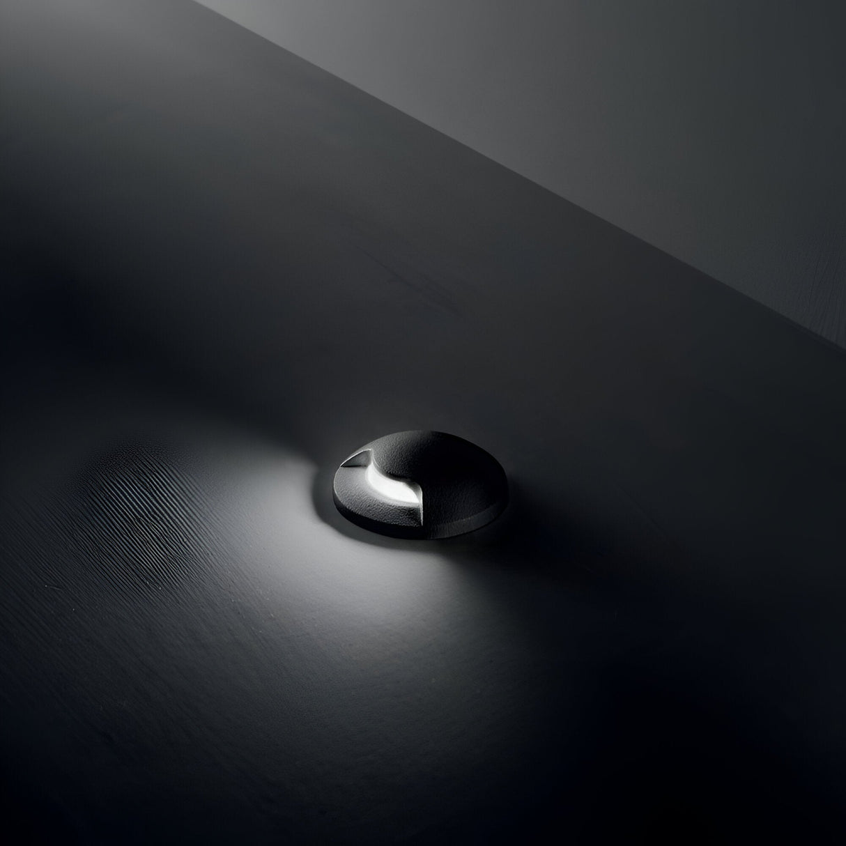 The Solarium Outdoor Drive Over Recessed Light One-Sided 3000K in black is positioned on a smooth, dark surface, featuring an IP67 rating. It projects a focused beam of light ideal for outdoor spaces, producing a delicate glow and casting gentle shadows around the area.
