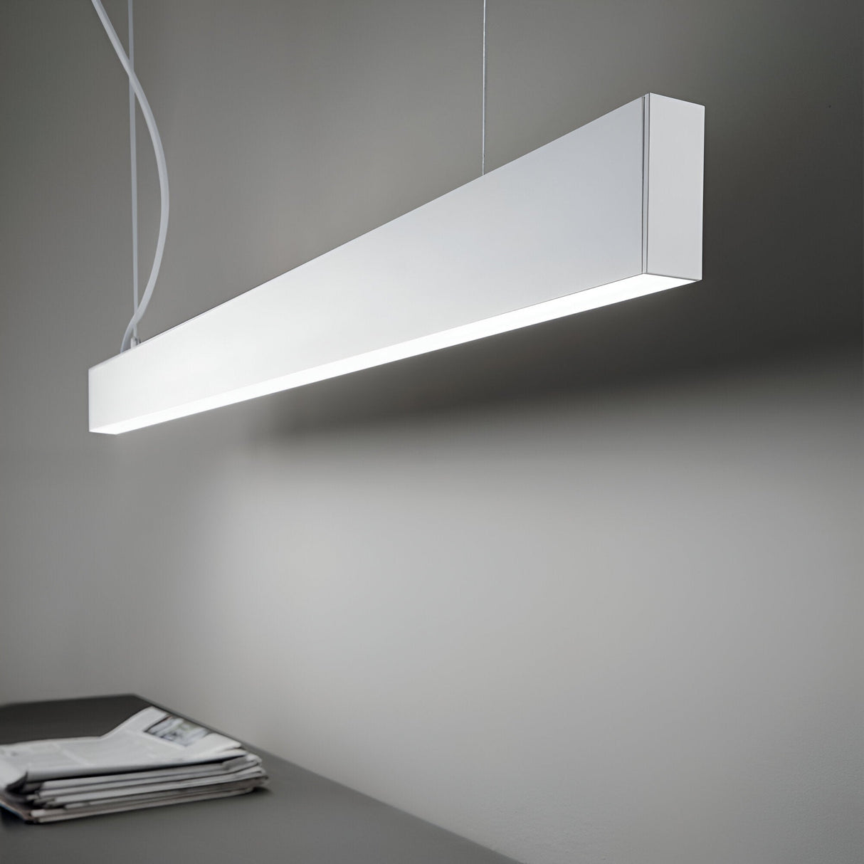 A sleek Ignitia-2 LED Suspension Light 36W 4000K in white with a rectangular design hangs from the ceiling, emitting a soft glow. Below it, a table holds a stack of newspapers. The room's contemporary interiors feature neutral colors and minimalist decor, enhanced by energy-efficient LED lighting.
