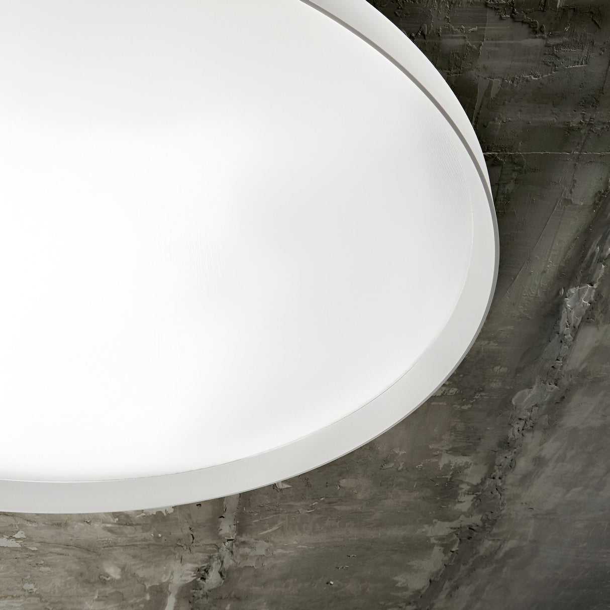 A portion of the Spark LED Flush Light 65W 4000K - White, 90cm is visible, mounted on a textured gray concrete ceiling. The large, round modern fixture emits a soft glow that highlights the rustic surface of the ceiling with its energy-efficient LED technology.