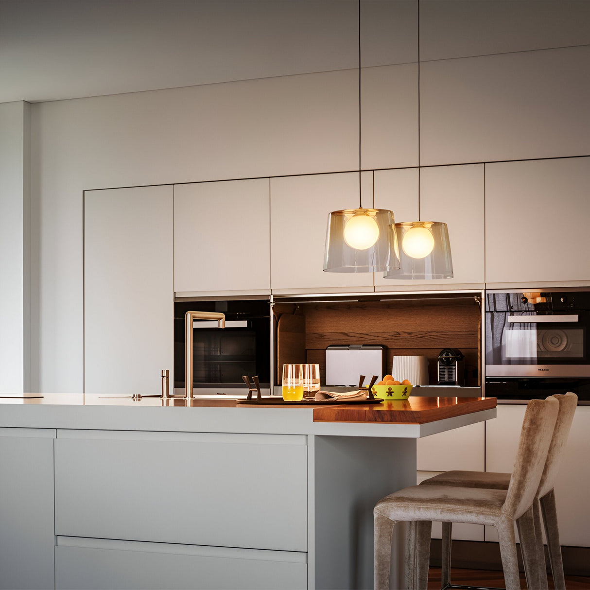 Modern kitchen with sleek white cabinets and stainless steel fixtures. A wooden bar extends from the countertop with stools underneath. Two Zenith 1 Light Glass Pendants in Brass hang above, illuminating the area. A glass of orange juice and fruit are on the counter, enhancing the contemporary style lighting.