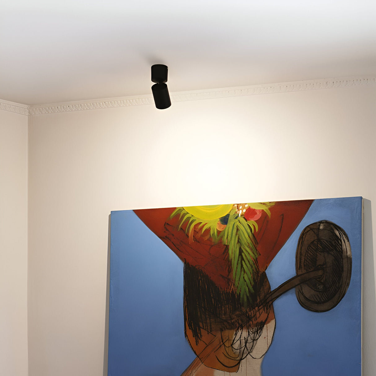 A modern art painting with abstract shapes and vibrant colors is hung on a beige wall. An Inlight Single Spotlight - Black, featuring adjustable diffusers on the ceiling, illuminates the artwork and enhances its allure under the modern lighting.
