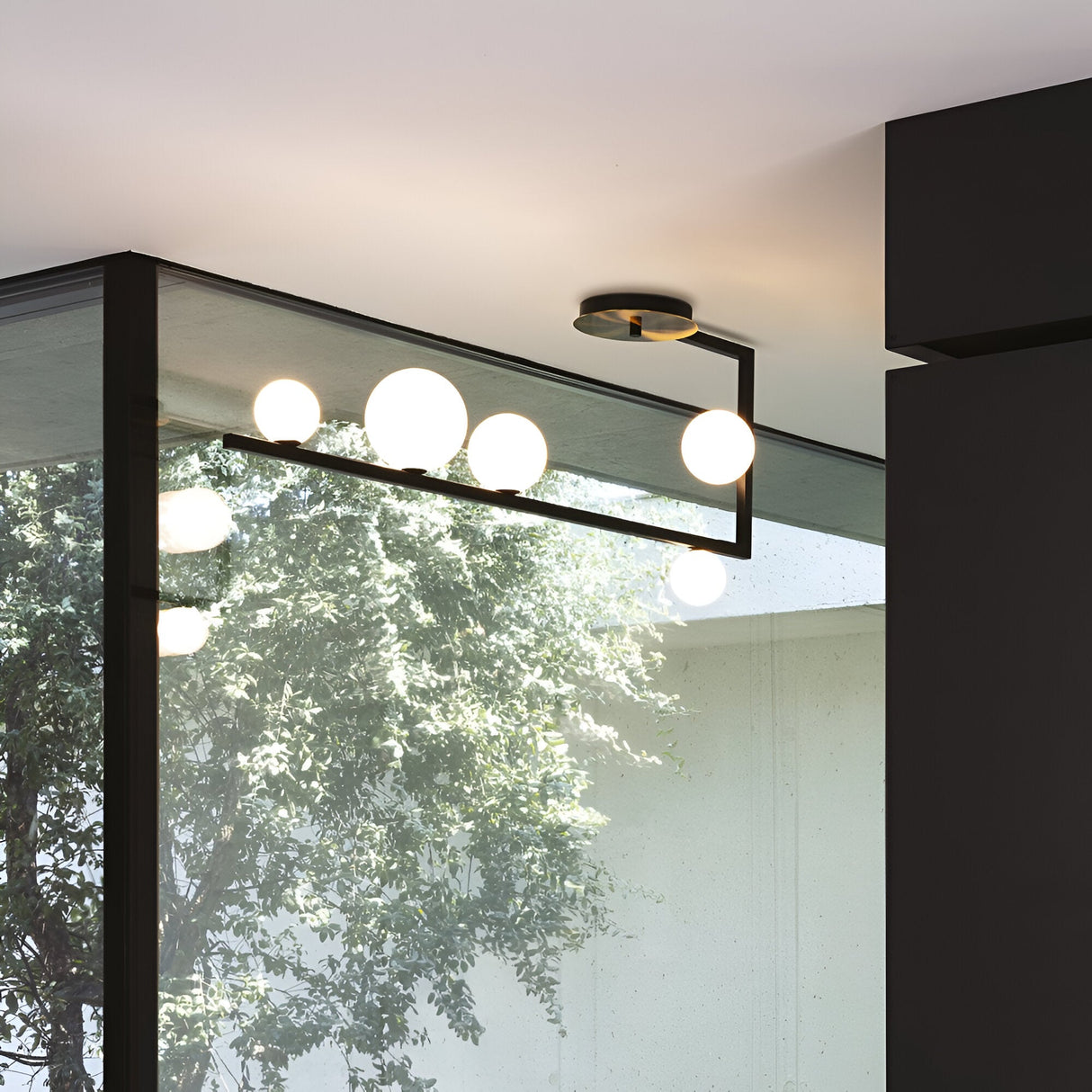 The Ethereal 5 Light Semi Flush - Black is a modern lighting fixture with a sleek black metal construction featuring spherical bulbs arranged on a geometric frame. Its minimalist design beautifully complements the ambiance of a room, where large windows reflect light and highlight the silhouette of a leafy tree outside.