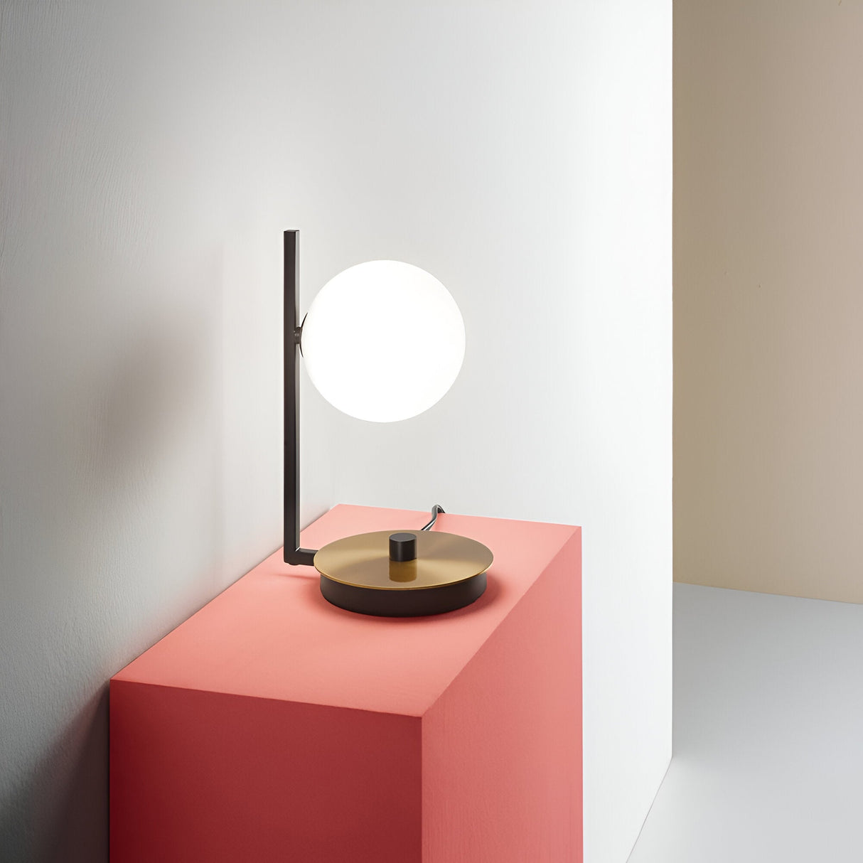 The Ethereal Table Lamp - White features a glowing round bulb resting on a sleek black stem with a metal finish base. It is positioned on a red-brown pedestal, surrounded by white and pale beige walls, resulting in a minimalist design aesthetic.
