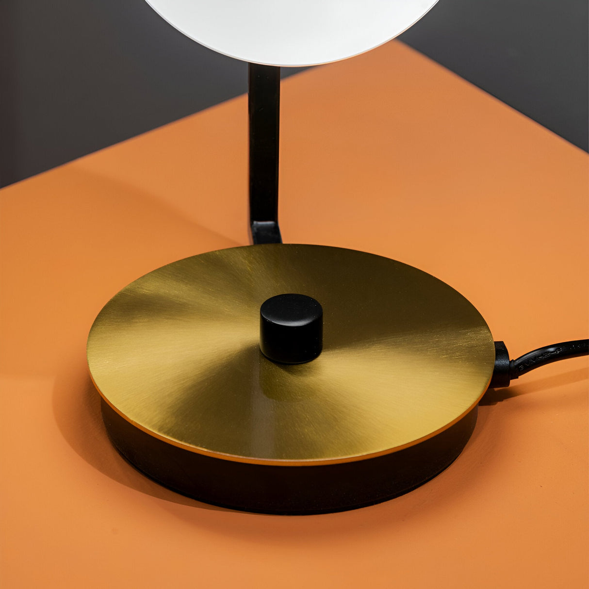 A close-up of the Ethereal Table Lamp - White on an orange surface, showcasing a round, gold metallic base with a sleek metal finish. The base features a small black knob in the center and has a black power cord extending from it, exemplifying a modern and minimalist design.