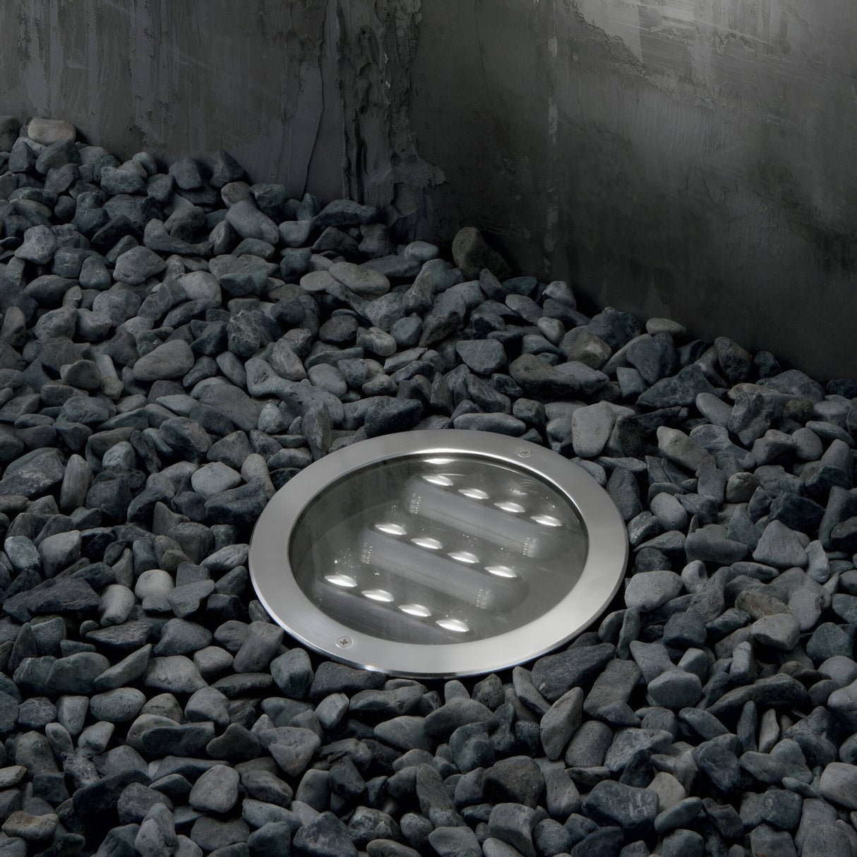 The Zephyra Outdoor LED Round Recessed Ground Light - 20W, 3000K, Grey is embedded in a circular metal casing and set among a bed of smooth gray pebbles near a textured concrete wall. With its IP67 rating, this light is ideal for enhancing your outdoor space while withstanding the elements.