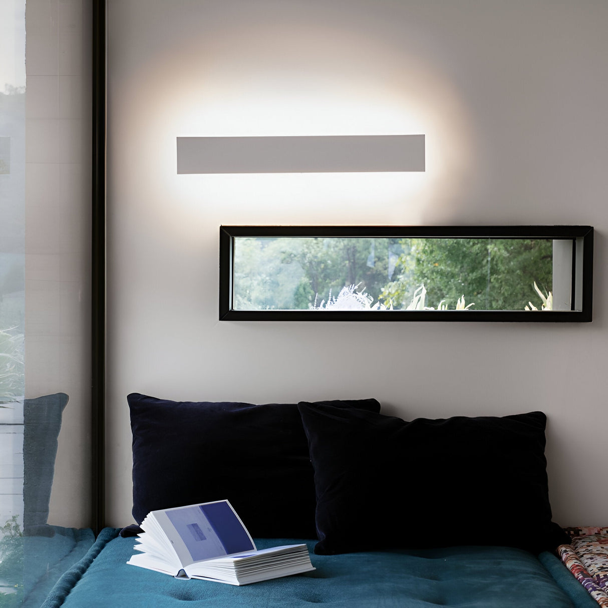 A cozy reading nook features a blue couch, accompanied by two navy pillows and an open book. A rectangular mirror adorns the wall, reflecting greenery from outside. The Flara LED Wall Light 23W 4000K in White, measuring 53cm, casts a warm glow above the mirror, enhancing the energy efficiency of this inviting space.