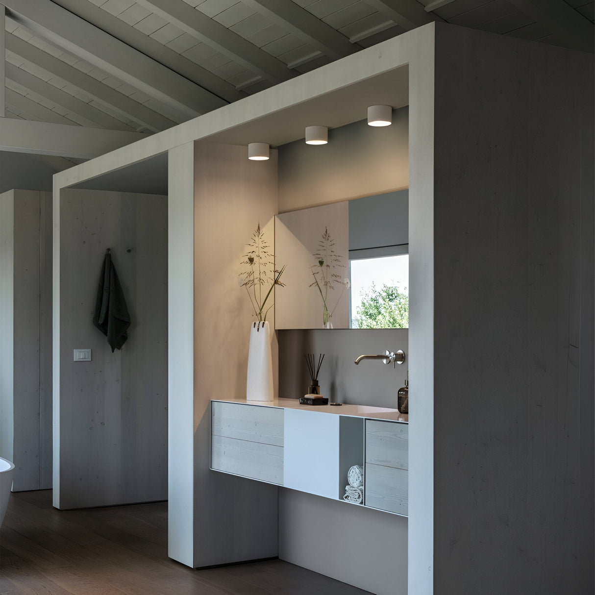 A modern bathroom with a minimalist design features a wall-mounted sink and a mirrored cabinet. The space is illuminated by three Orbis Round Downlights in white, complemented by decorative vases with plants and a small clock on the counter. Light wood and white tones dominate the atmosphere.