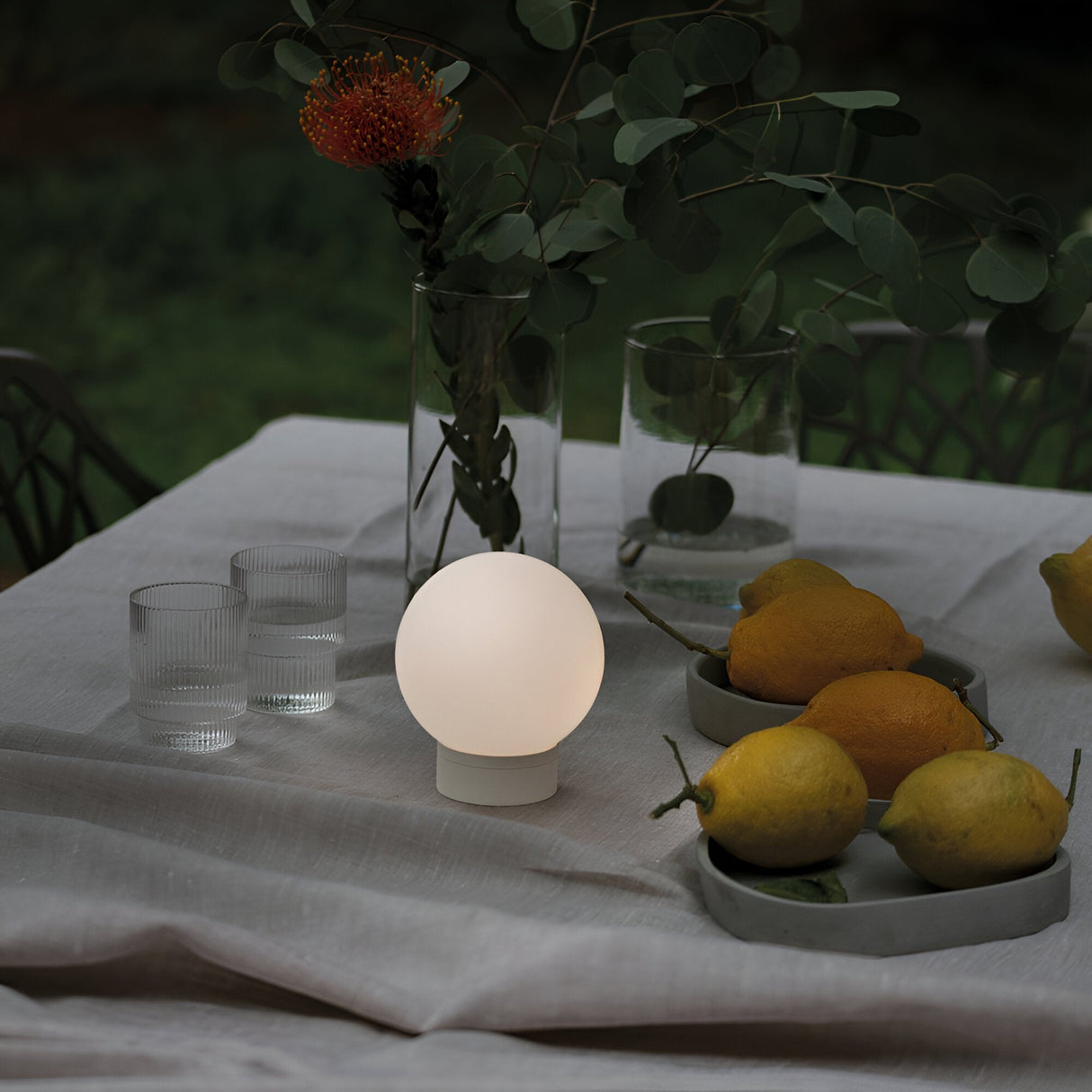 A dimly lit outdoor table, adorned with a white cloth, features the Beam Outdoor Table Lamp in White, illuminating the scene with its radiant LED bulb inside a spherical design. Two clear glasses, a vase holding green leaves and a red flower, along with a plate of lemons elevate the minimalist style. The backdrop consists of a softly blurred natural setting.