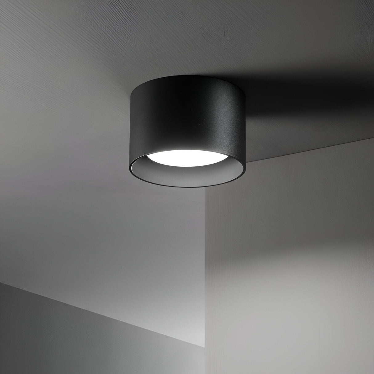The Orbis Round Downlight - Black, a modern cylindrical ceiling LED light fixture, brightens up a dimly lit room. Its downlight casts a soft, focused glow on the surrounding grey ceiling and walls, cultivating a minimalist and elegant atmosphere.