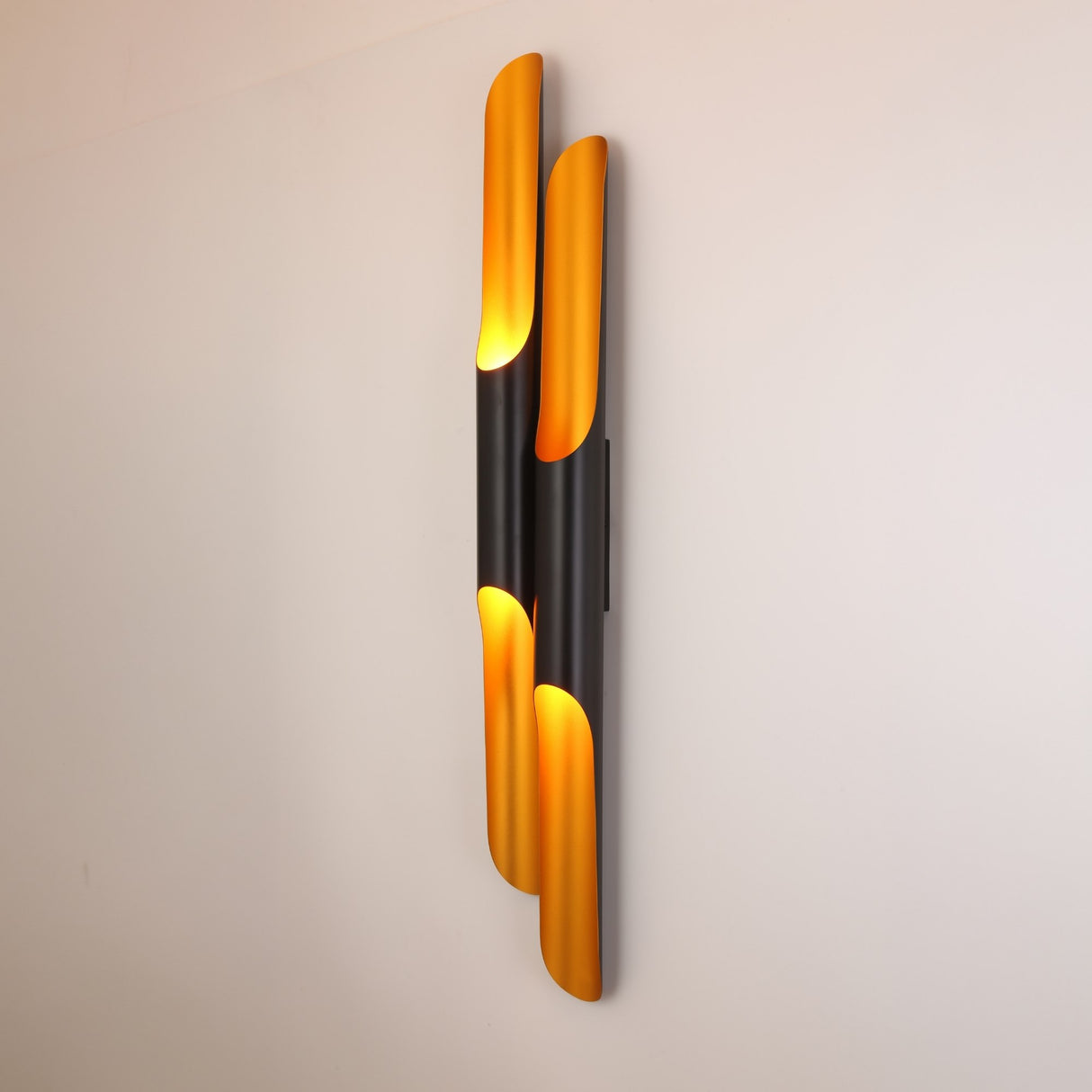 The Stealth 4 Light Wall Light in brass and black features elongated curved panels, casting a warm yellow glow that adds elegant, contemporary style and striking contrast to any plain white wall.