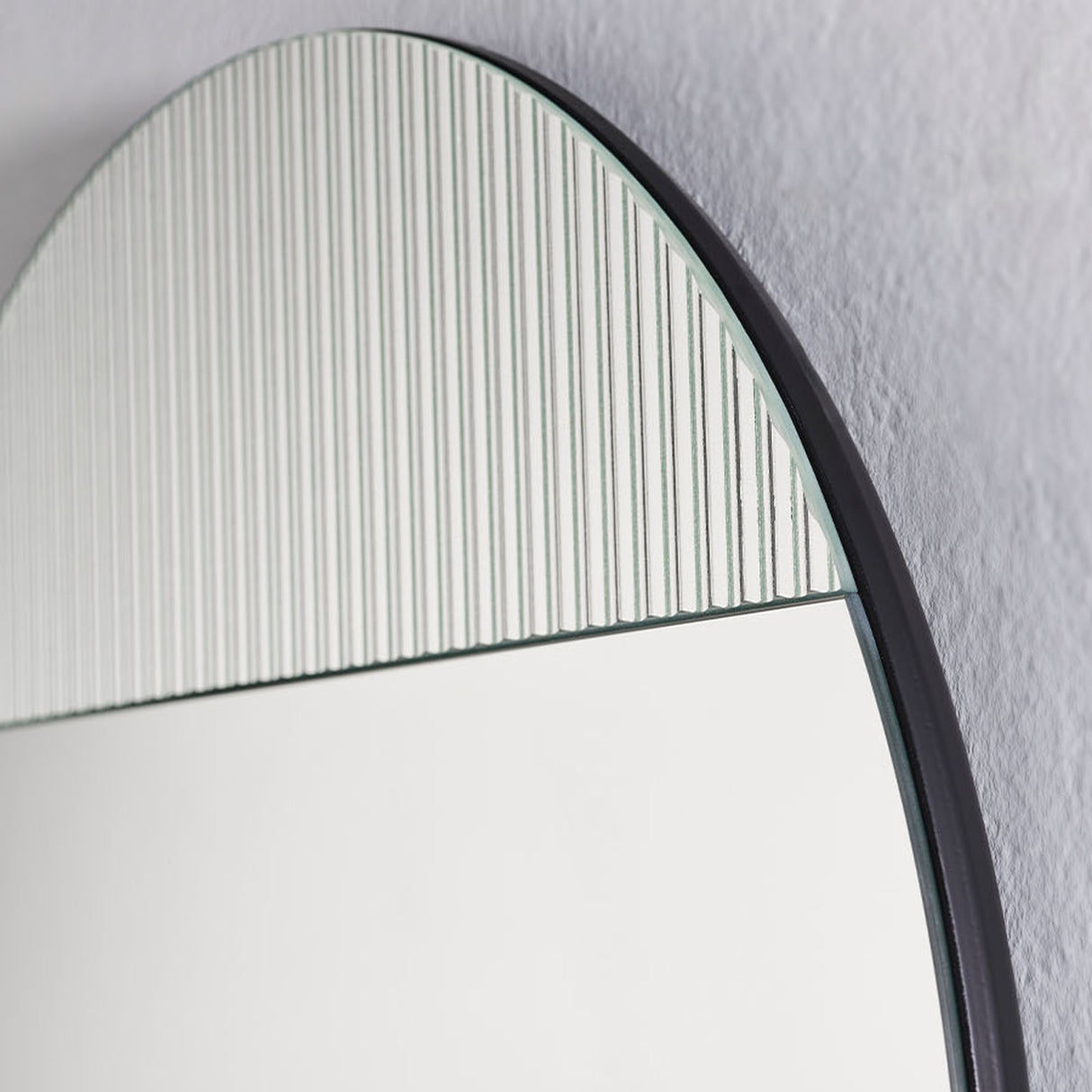 Close-up of Cord Flutes round wall mirror, 58cm, with reeded glass ribbed upper section meeting plain mirror below, thin black frame, on a light grey wall.