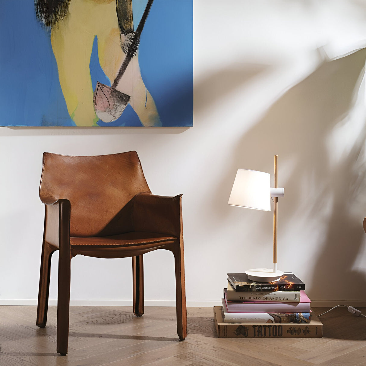 A stylish room corner features a modern brown leather chair on a light wooden floor. Resting on a stack of books is the Twine Wood Table Lamp in white, offering sleek adjustable lighting. Part of a colorful abstract painting graces the wall, enhancing the space's modern design.