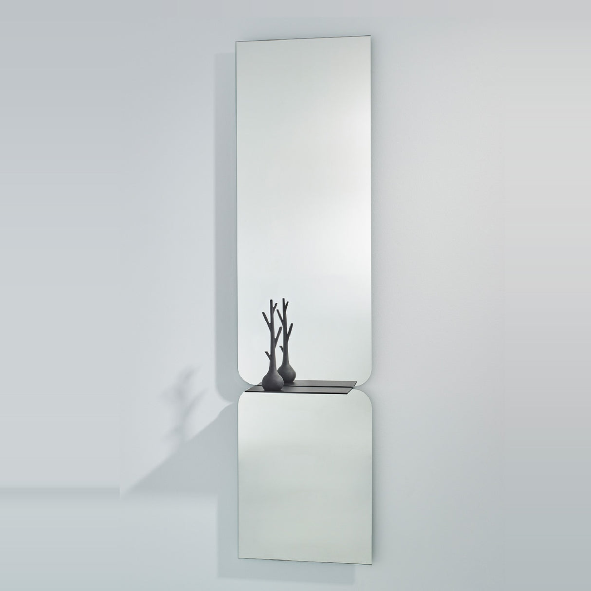Taille full length hall mirror, 180cm, frameless rectangular glass in two panes with rounded join and black metal shelf, shown on a plain light grey wall.
